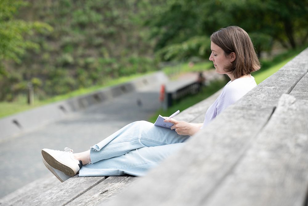 Eine Frau sitzt im Freien auf einer Betontreppe und schreibt in ein Notizbuch.
