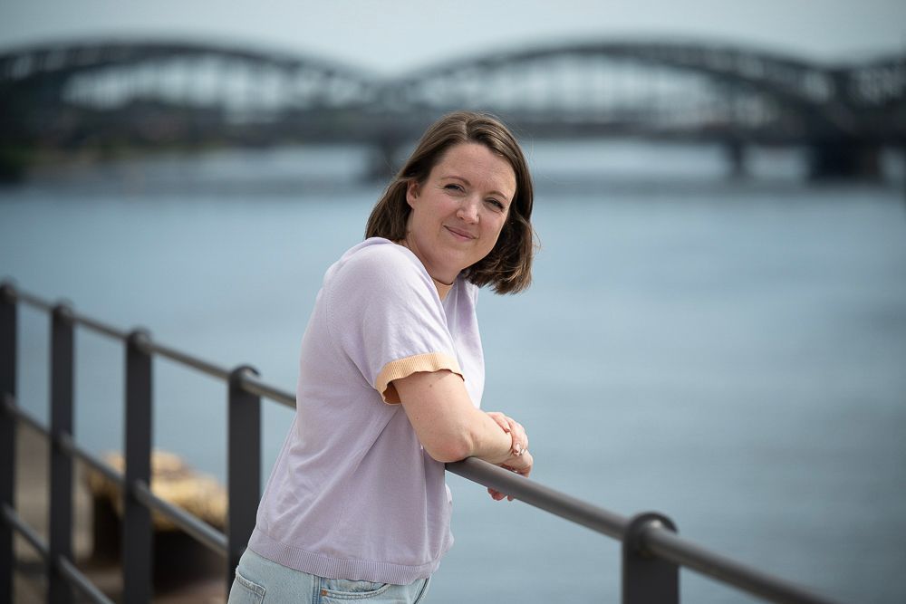 Frau lehnt lächelnd an einem Geländer; im Hintergrund sind eine Brücke und Wasser zu sehen.