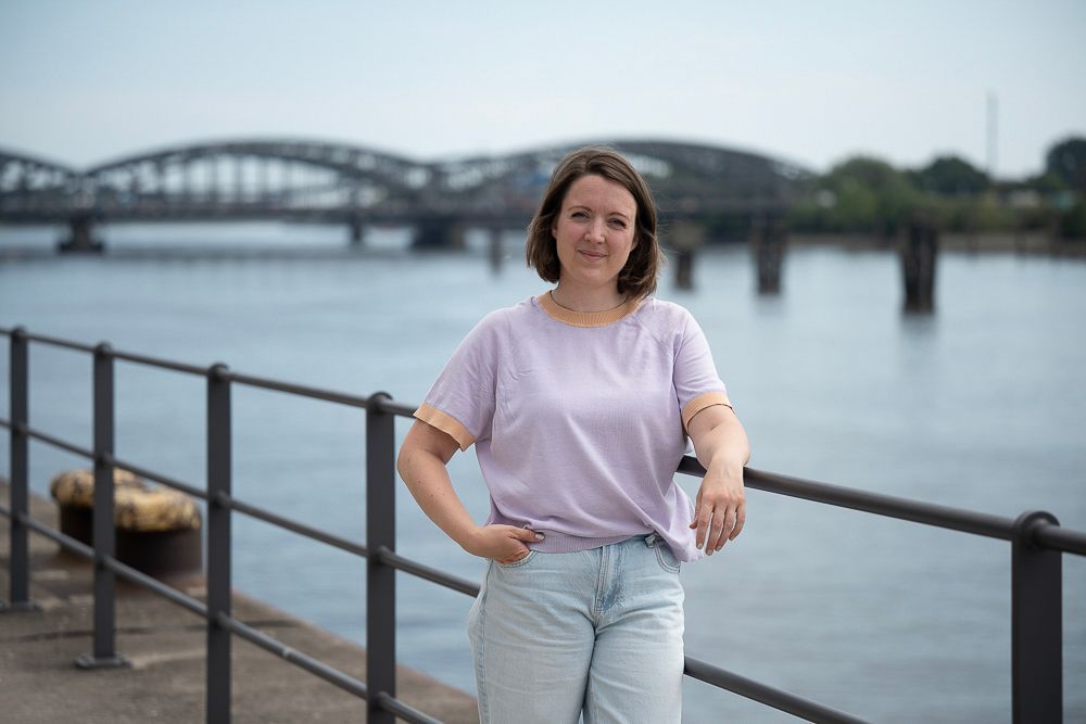 Eine Frau in lavendelfarbenem Hemd und Jeans steht an einem Fluss, im Hintergrund ist eine Brücke zu sehen.