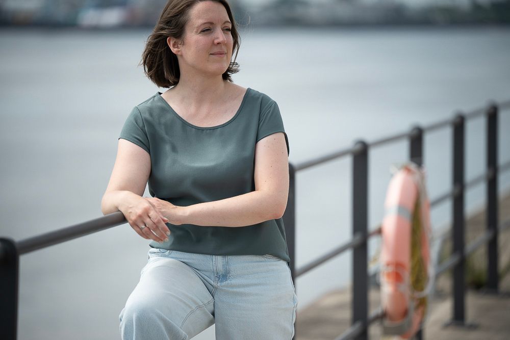 Eine Frau lehnt am Geländer und blickt aufs Wasser. Sie trägt ein grünes Hemd und Jeans. In der Nähe liegt ein Rettungsring.
