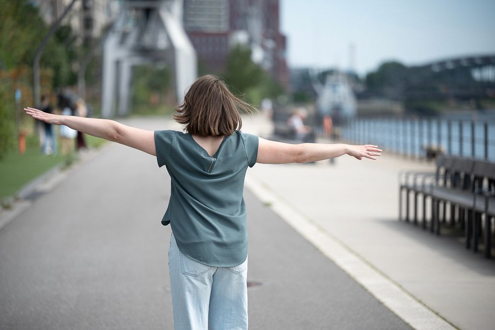 Eine Person mit ausgestreckten Armen steht auf einem gepflasterten Weg in der Nähe von Wasser; im Hintergrund sind moderne Gebäude zu sehen.