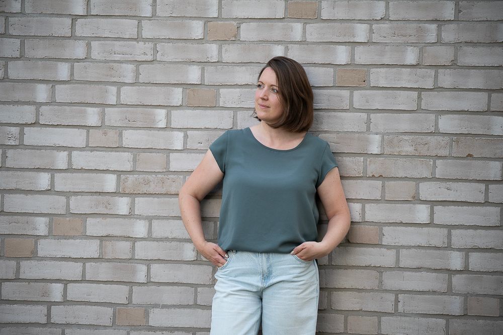 Eine Frau in grünem Top und Jeans lehnt an einer Backsteinmauer und blickt zur Seite.