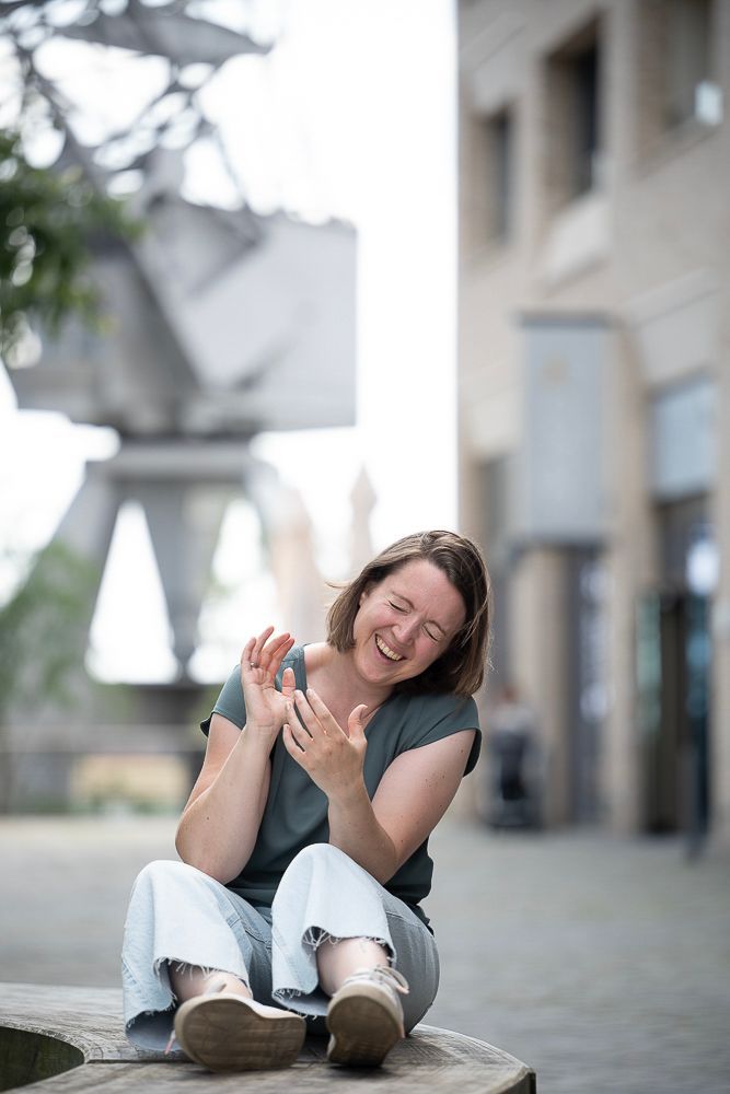 Frau sitzt im Freien, lacht und hebt die Hände. Sie trägt ein grünes Oberteil, eine helle Hose und beige Schuhe. Städtische Umgebung.