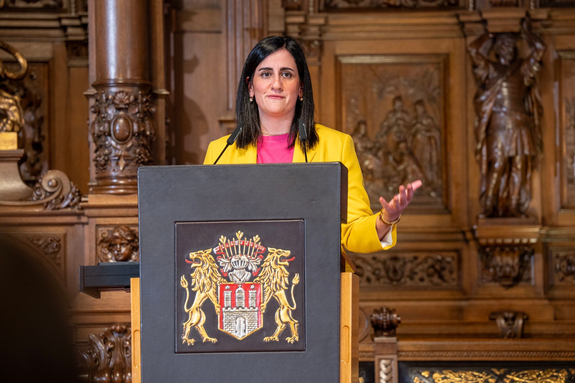 Eine Frau in einem gelben Blazer spricht an einem Podium mit Wappen in einem reich verzierten Holzzimmer.