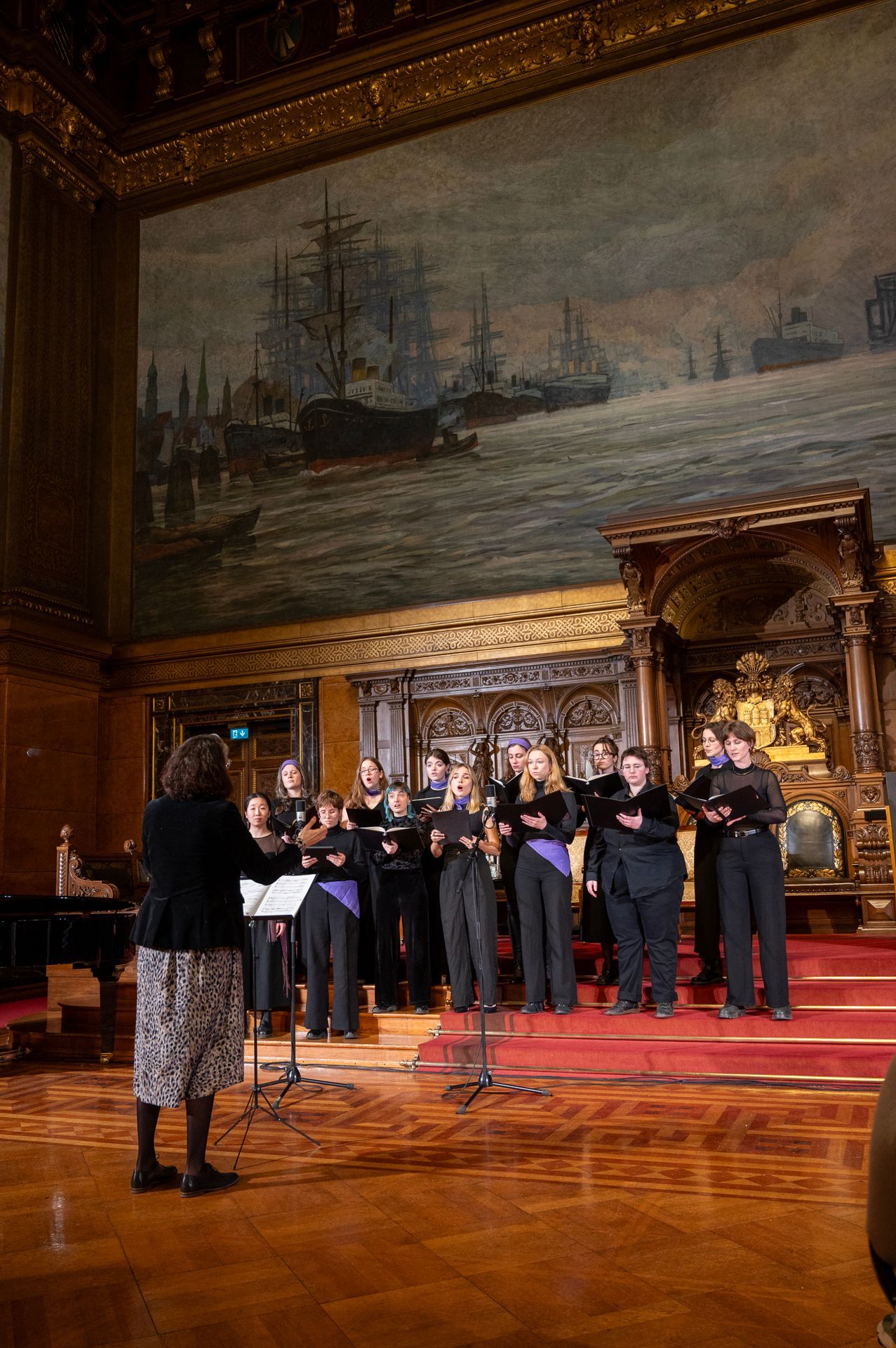 Ein Chor singt auf der Bühne, der Dirigent steht ihnen gegenüber. Hinter ihnen hängt ein Wandgemälde mit Schiffen. Roter Teppich.