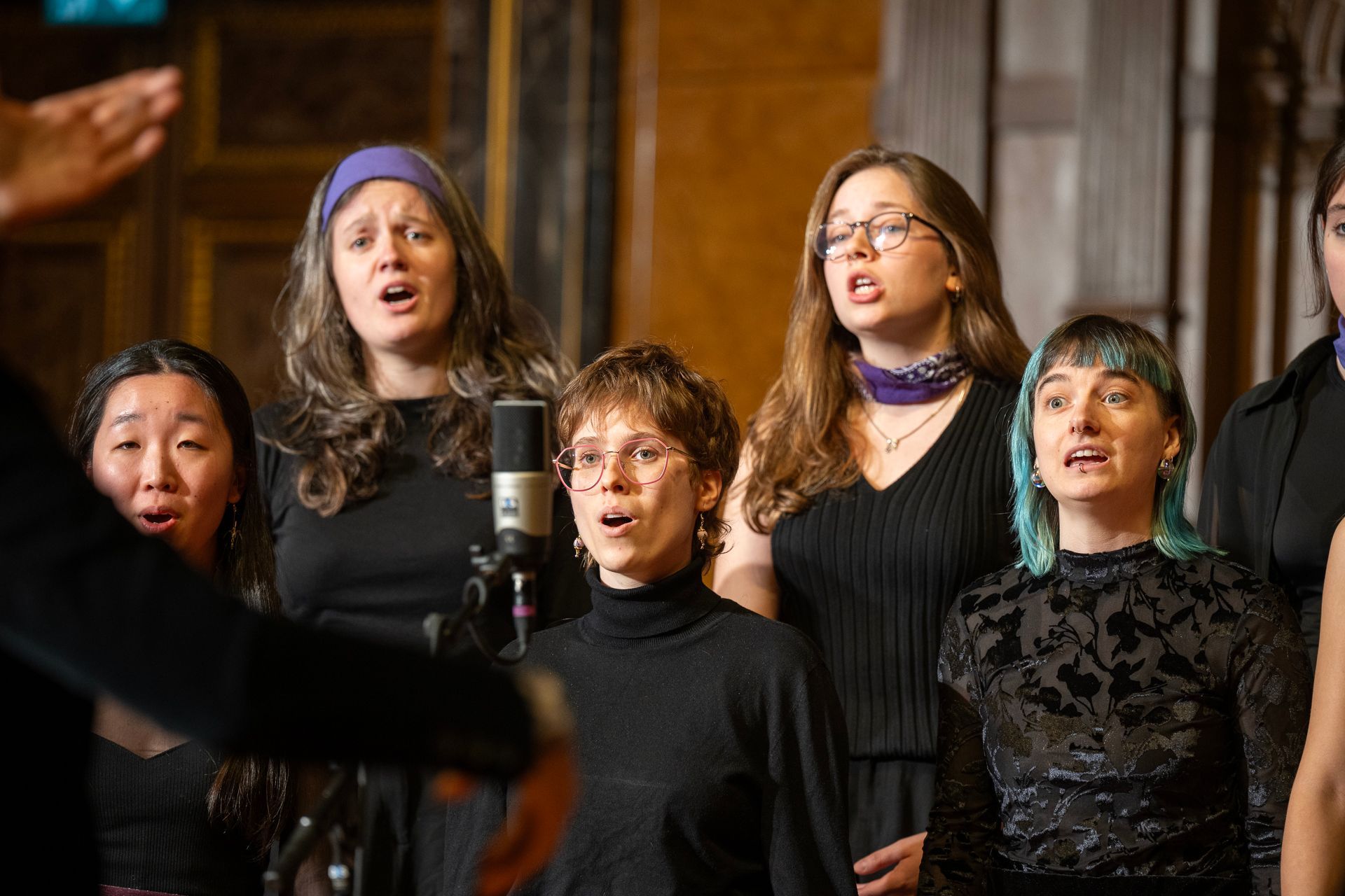 Ein Chor singt in einem Raum; einige blicken nach oben, andere haben den Mund offen. Ein Dirigent gibt Gesten.