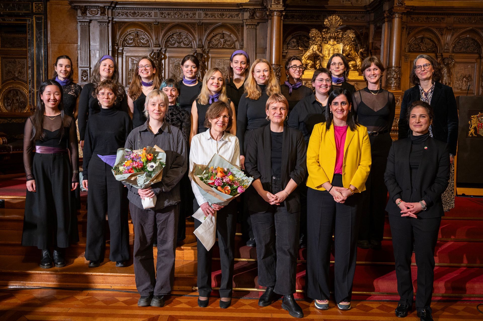 Gruppenfoto eines Chors; Frauen in schwarzen Outfits und violetten Schals, einige halten Blumensträuße, posieren auf einem Holzboden. Aufwendig verzierter Hintergrund.