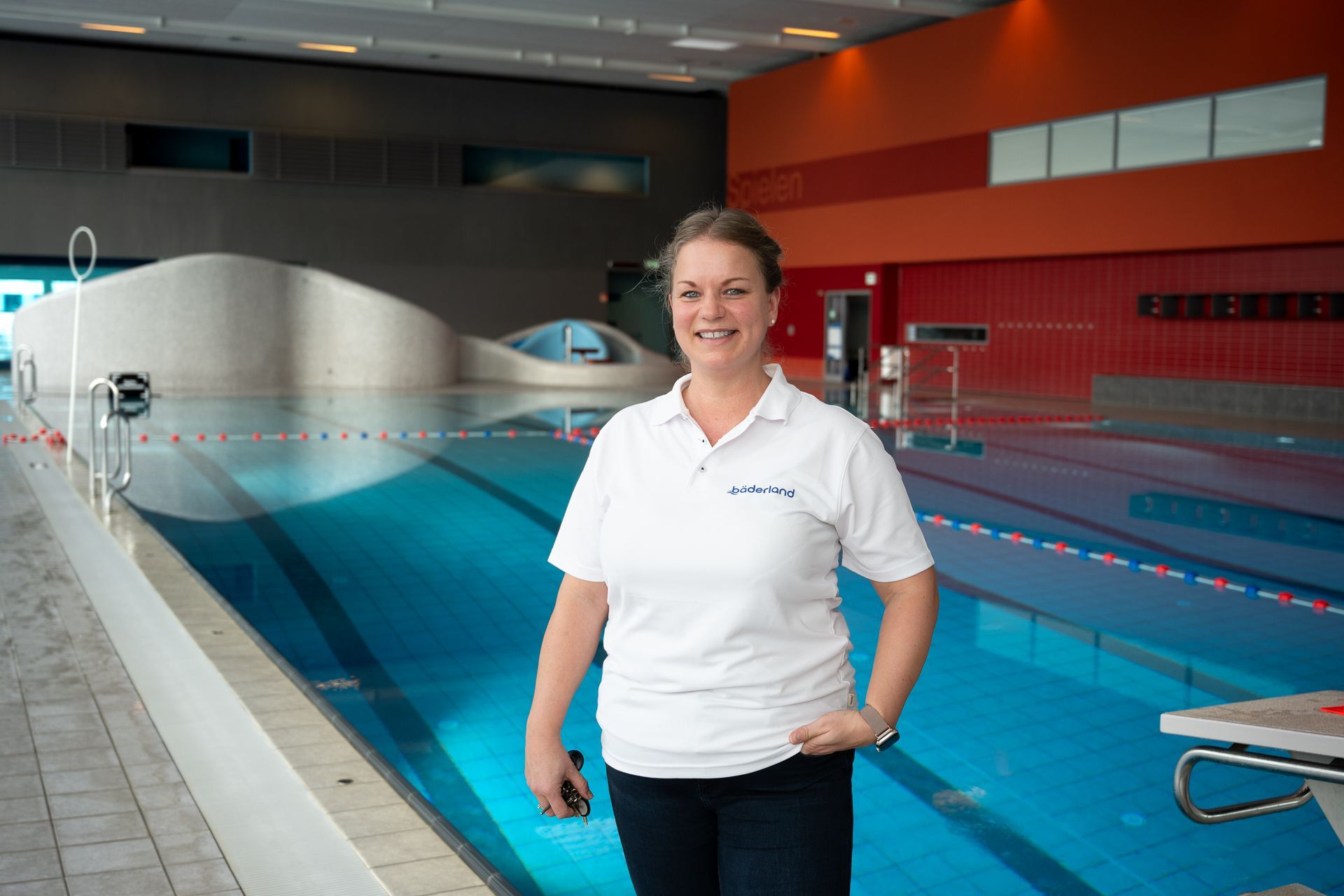 Eine Frau in einem weißen Poloshirt steht an einem Swimmingpool. Im Hintergrund ist eine orange-rote Wand zu sehen.