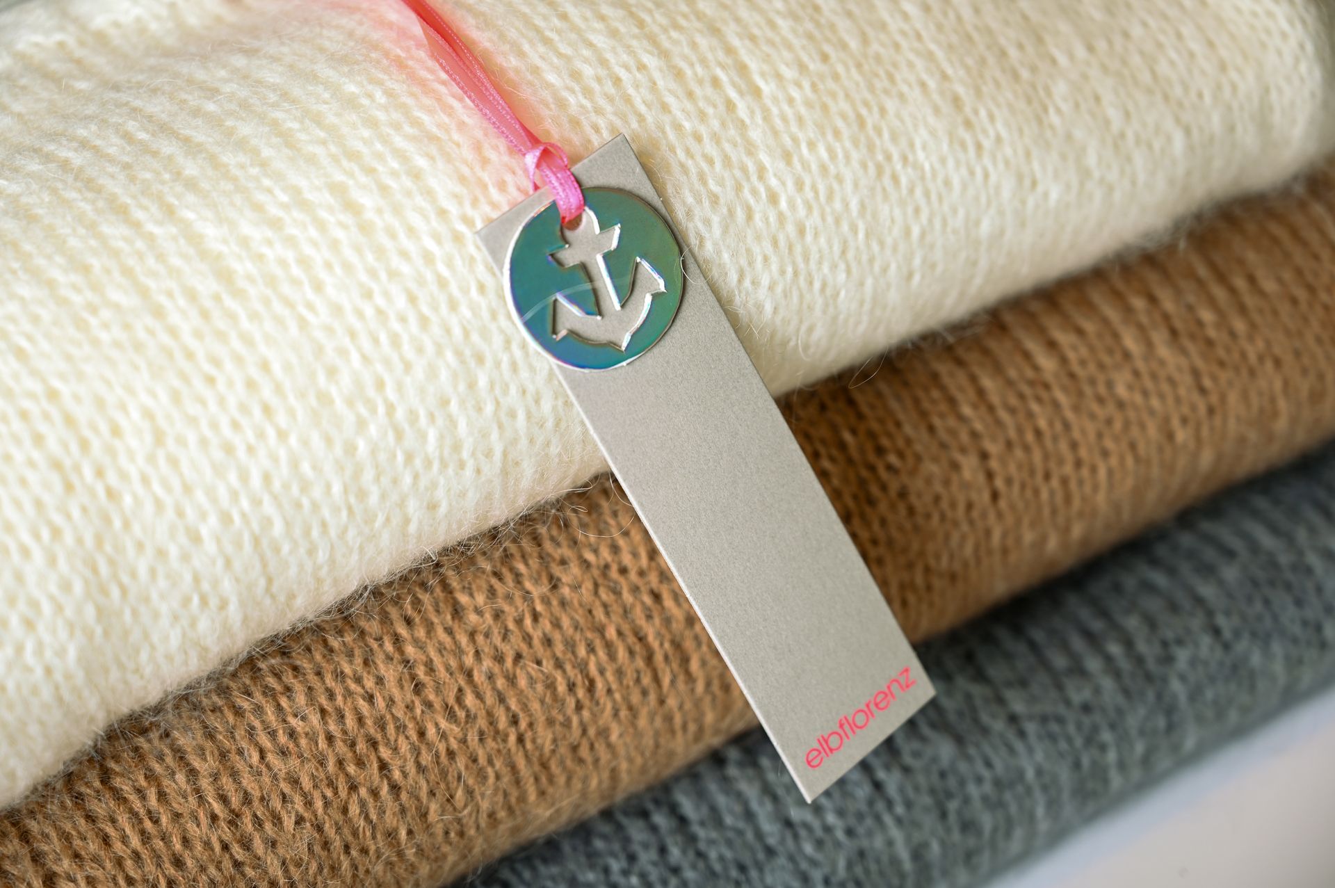 Stapel gefalteter Pullover in Weiß, Braun und Grau mit einem Etikett, das ein Ankersymbol zeigt.