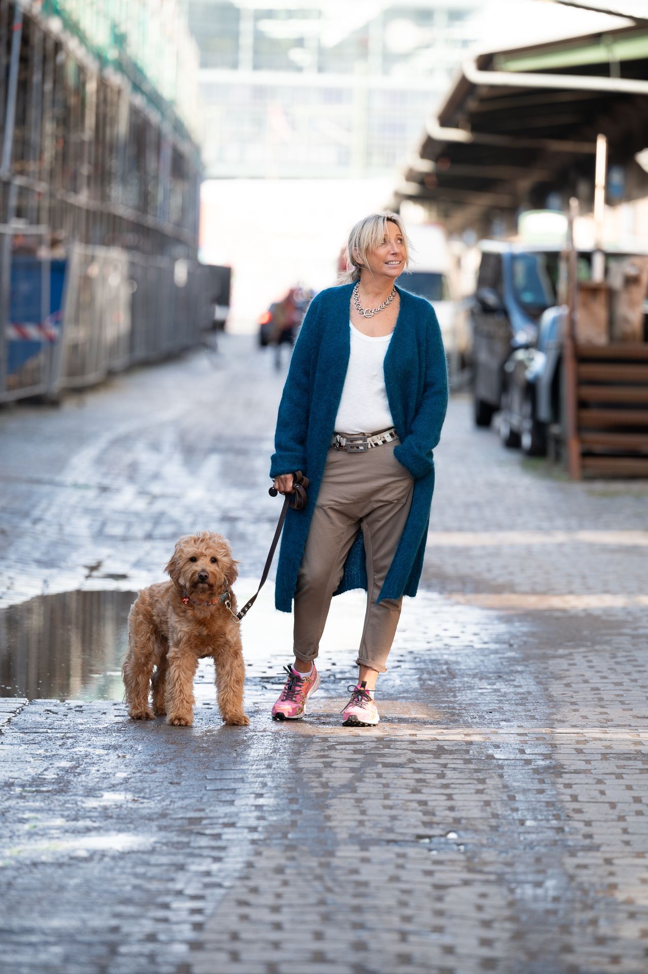 Frau, die einen Golden Doodle an der Leine im Freien ausführt, trägt eine lange blaue Strickjacke und lächelt.