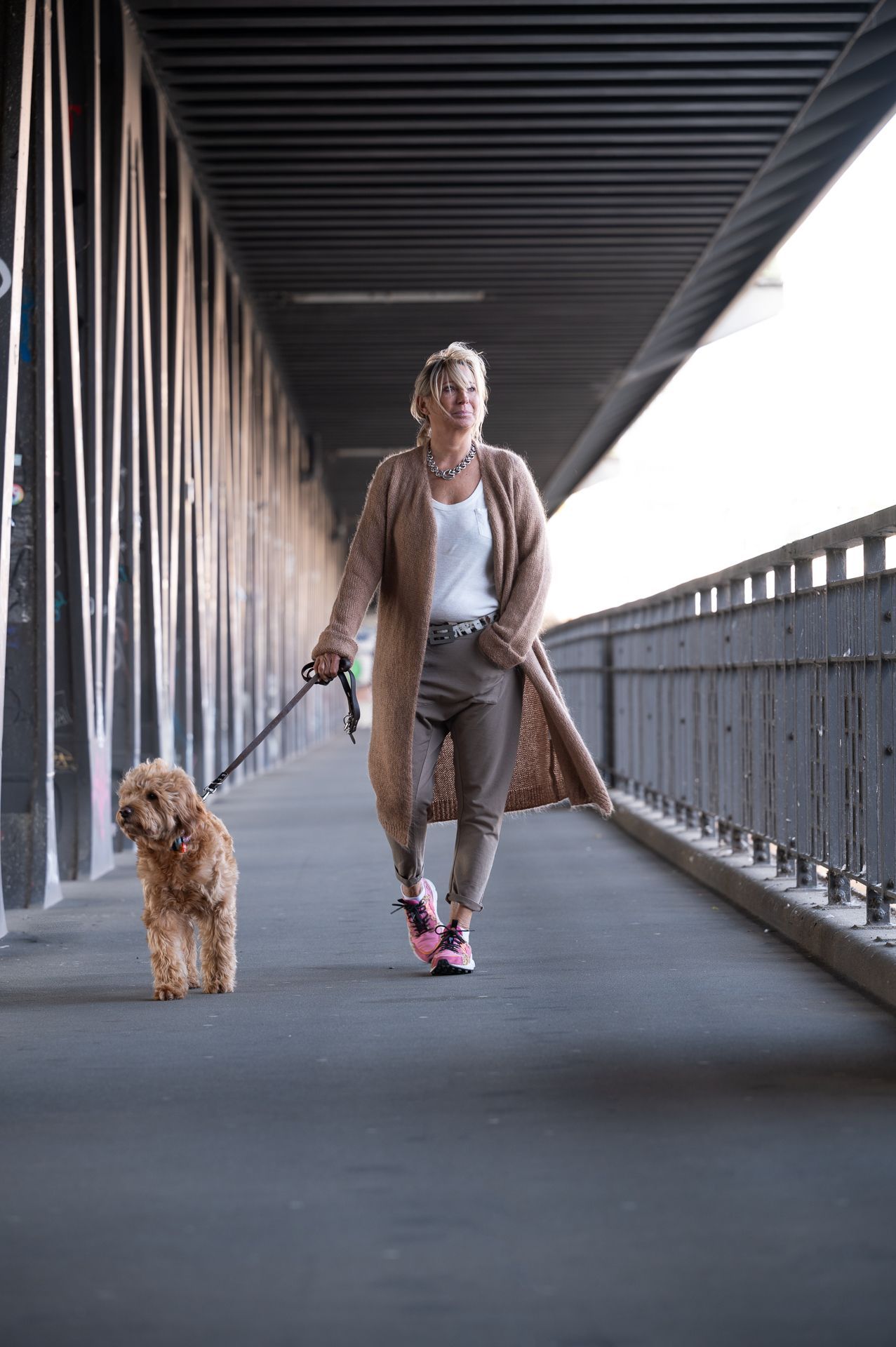 Eine Frau führt ihren Hund auf einer Brücke spazieren. Sie trägt eine lange Strickjacke, eine Hose und Turnschuhe. Der Hund ist an der Leine.