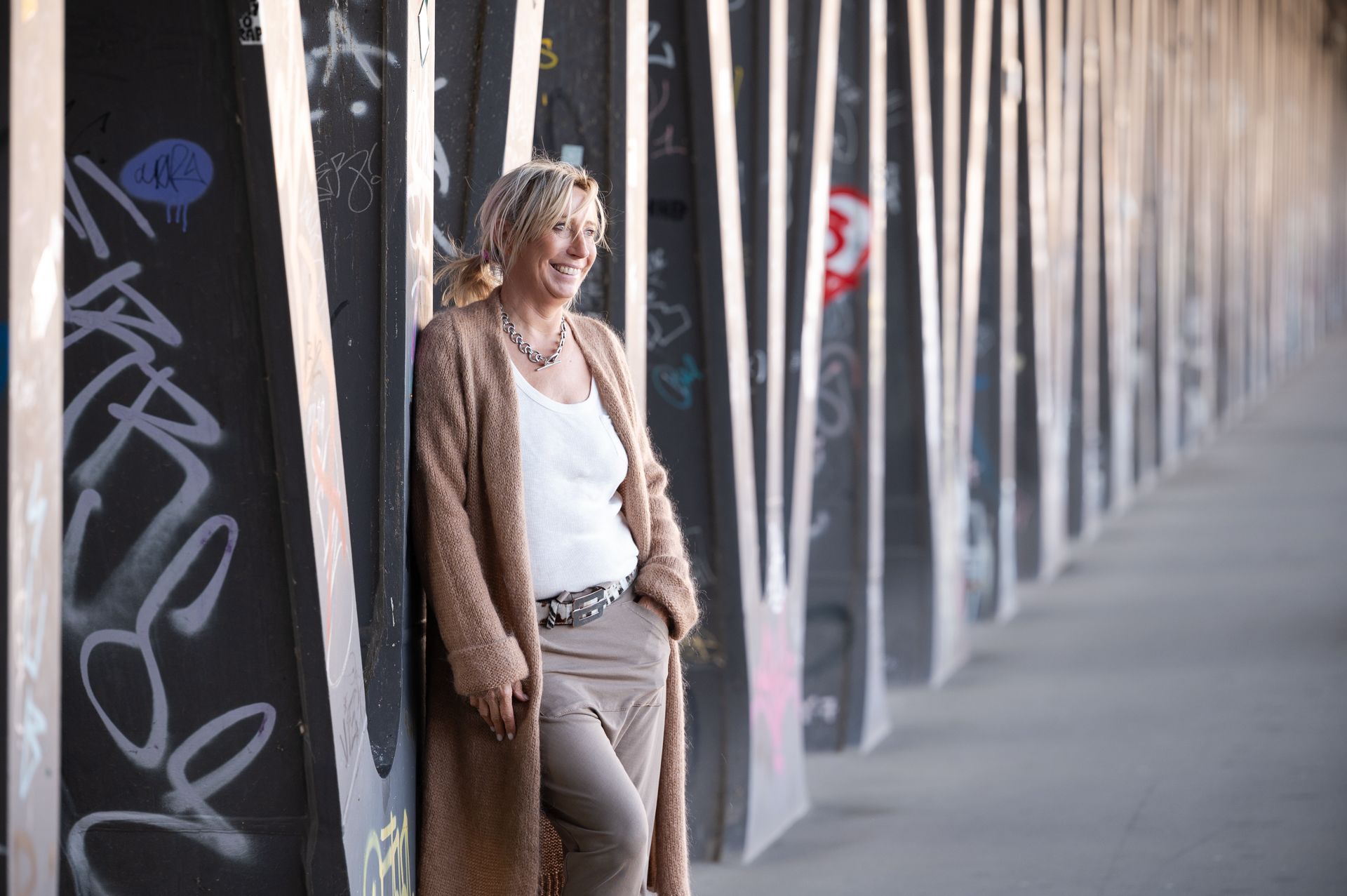 Eine Frau in einem langen, hellbraunen Cardigan lehnt lächelnd an einer mit Graffiti bedeckten Betonmauer.