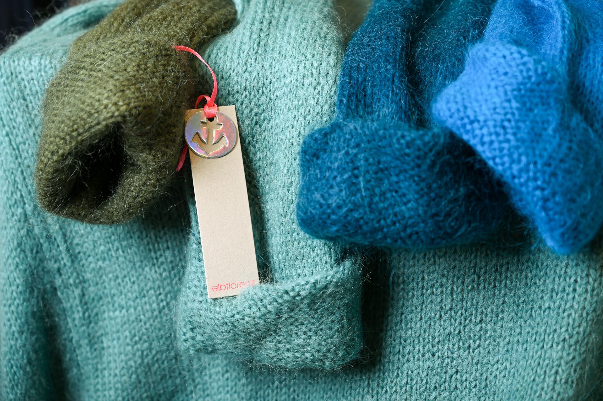 Nahaufnahme von drei Strickmützen: grün, türkis und blau. An der türkisfarbenen Mütze hängt ein Preisschild.