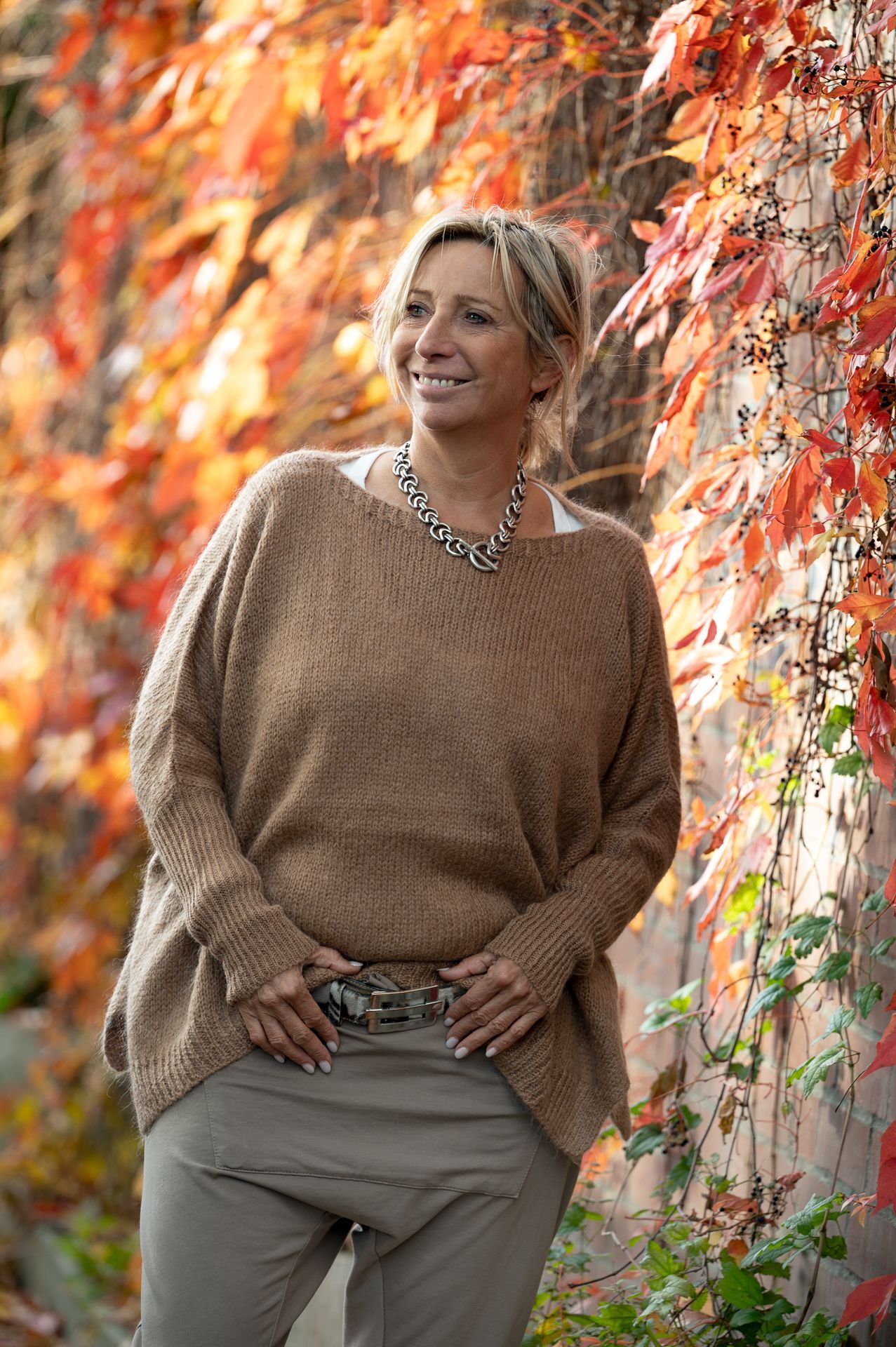 Eine Frau in braunem Pullover und Hose lächelt und steht neben roten Herbstblättern.