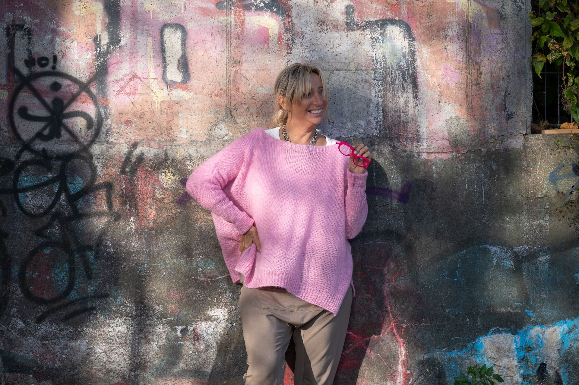 Frau in rosa Pullover posiert vor einer mit Graffiti bedeckten, von Sonnenlicht erhellten Wand.