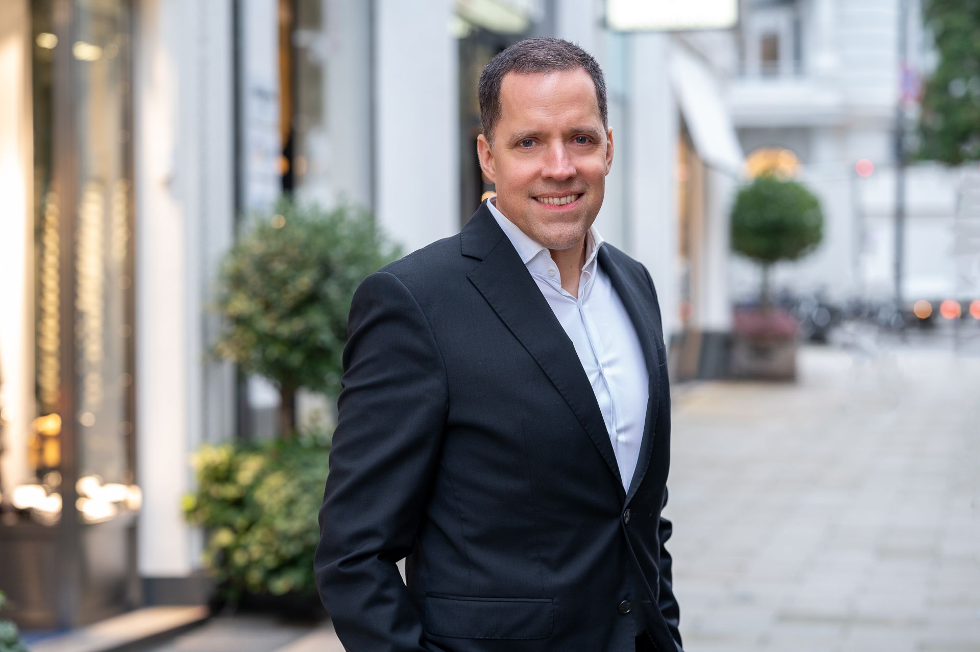 Ein Mann im Anzug lächelt, er steht auf einer Straße in der Stadt, im Hintergrund sind Geschäfte und Pflanzen zu sehen.