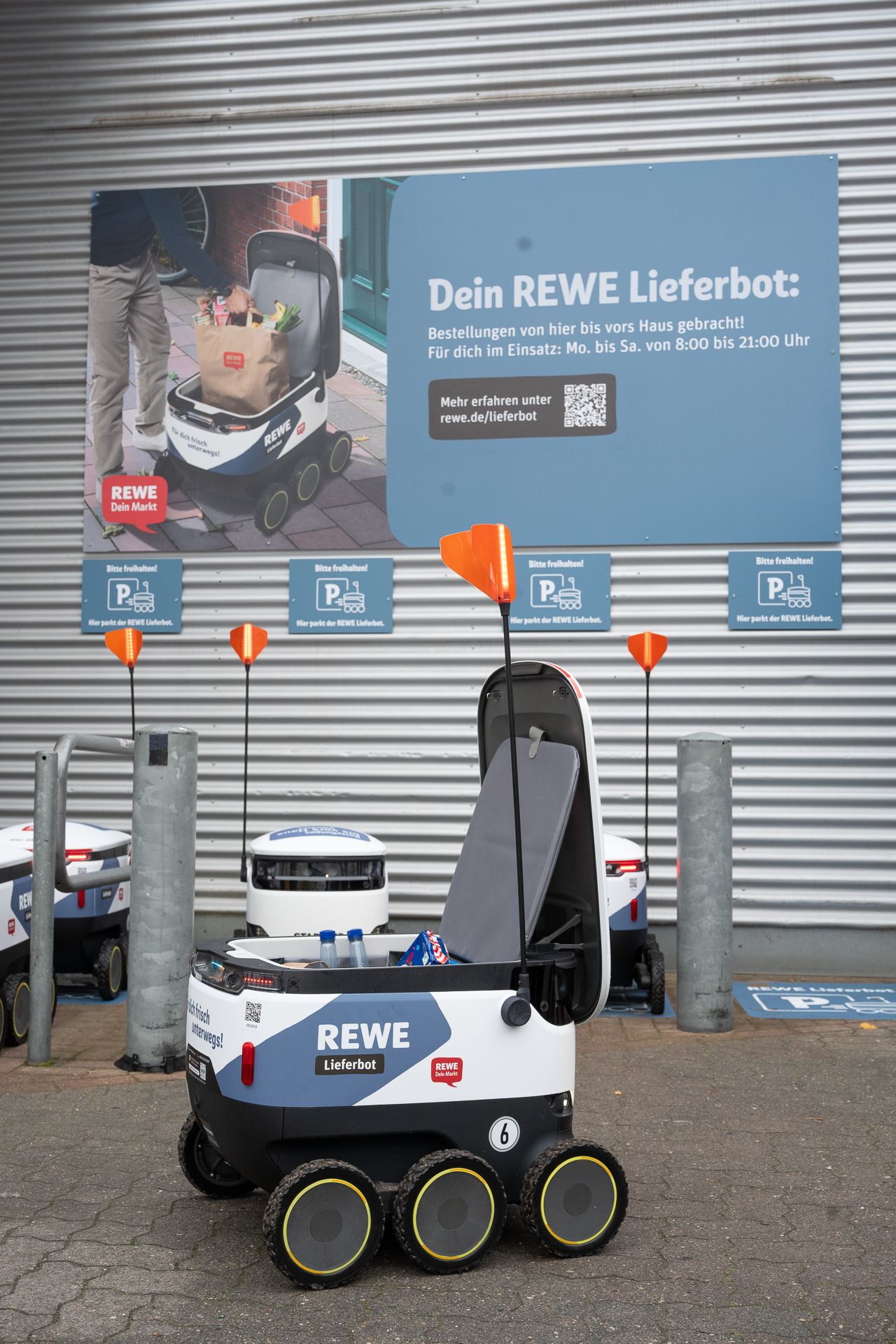 Ein REWE-Lieferroboter mit orangefarbener Flagge steht vor einem Geschäft mit einem Schild.