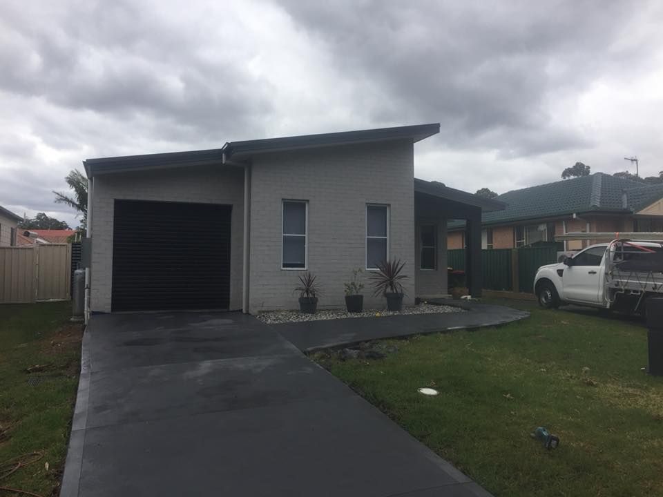 Small gray house with a black garage door, driveway, and a white truck parked beside it under cloudy skies