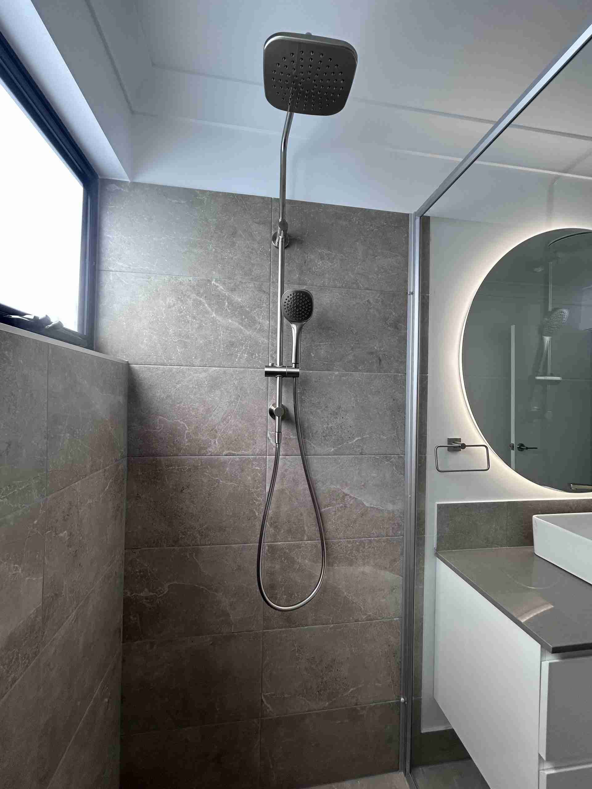Modern bathroom shower with gray tile, rainfall head, handheld sprayer, and round mirror sink vanity