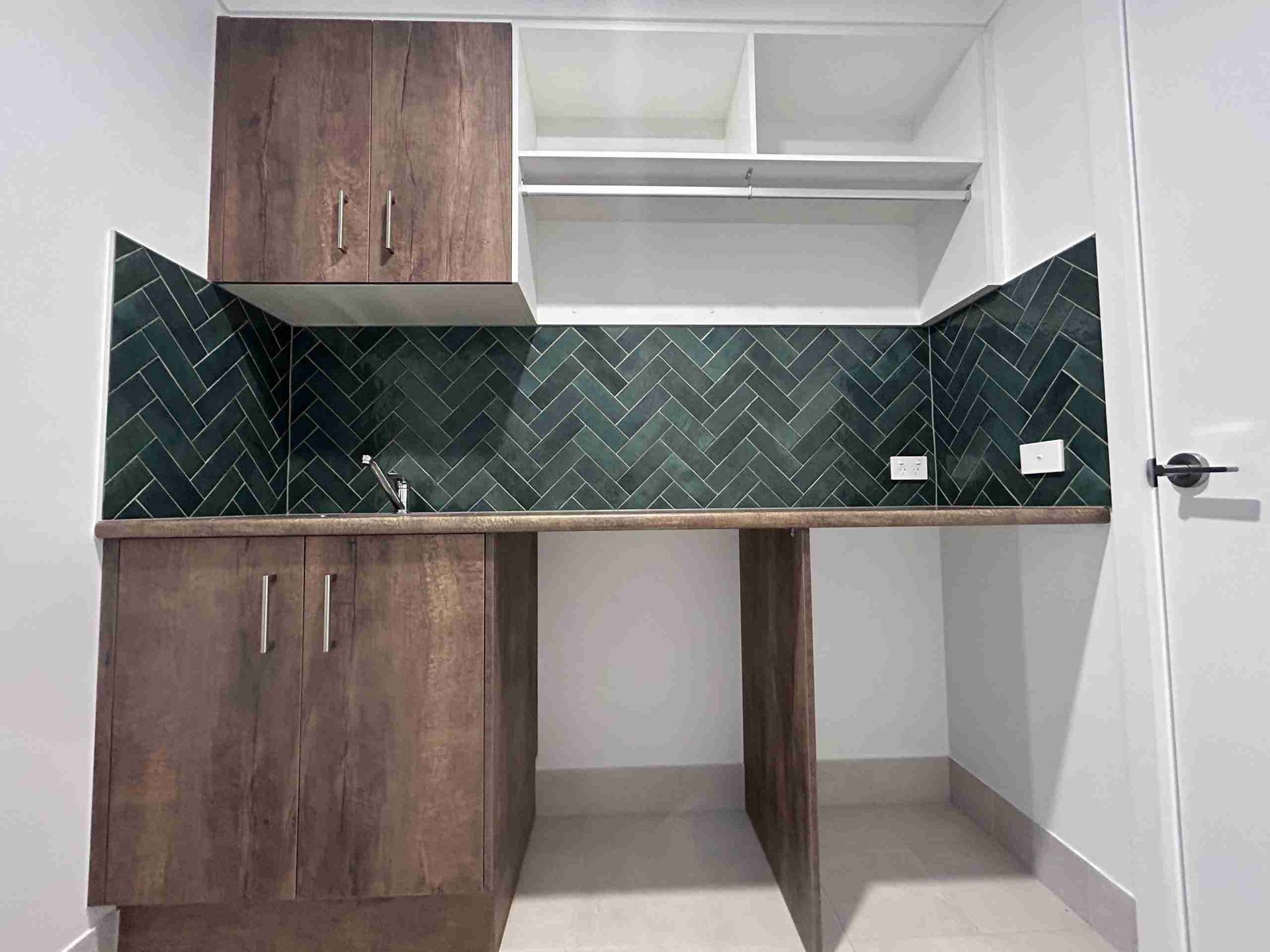 Small kitchen nook with dark wood cabinets, green tile backsplash, and a countertop under white shelves.