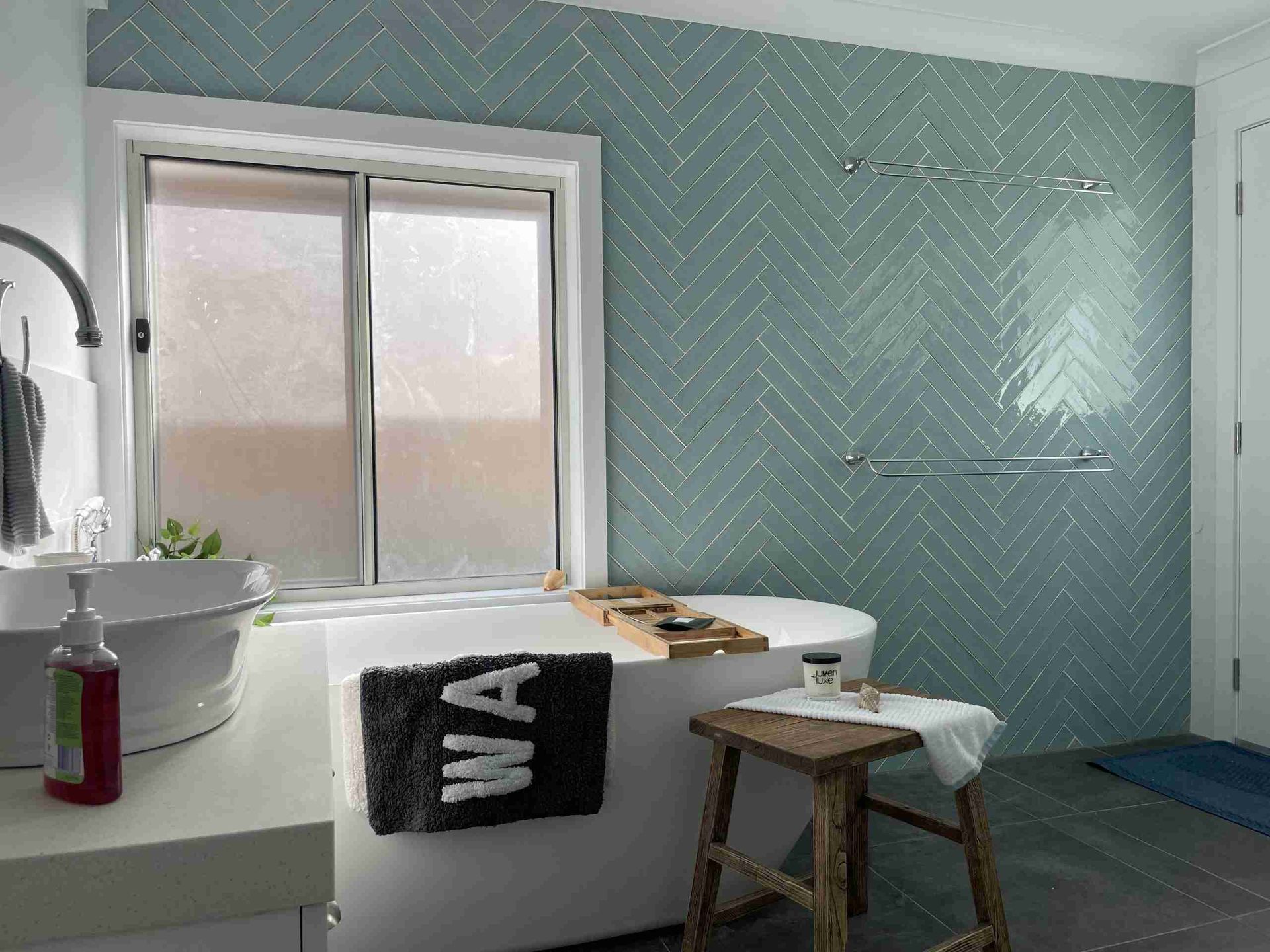 Bright bathroom with a tub, sink, window, herringbone blue wall, stool, and towels.