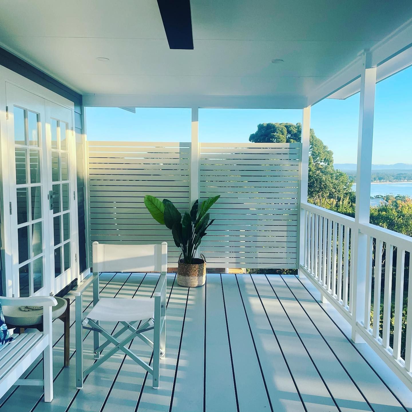 Sunlit porch with white railings, glass doors, a table and chairs, and a potted plant overlooking water.