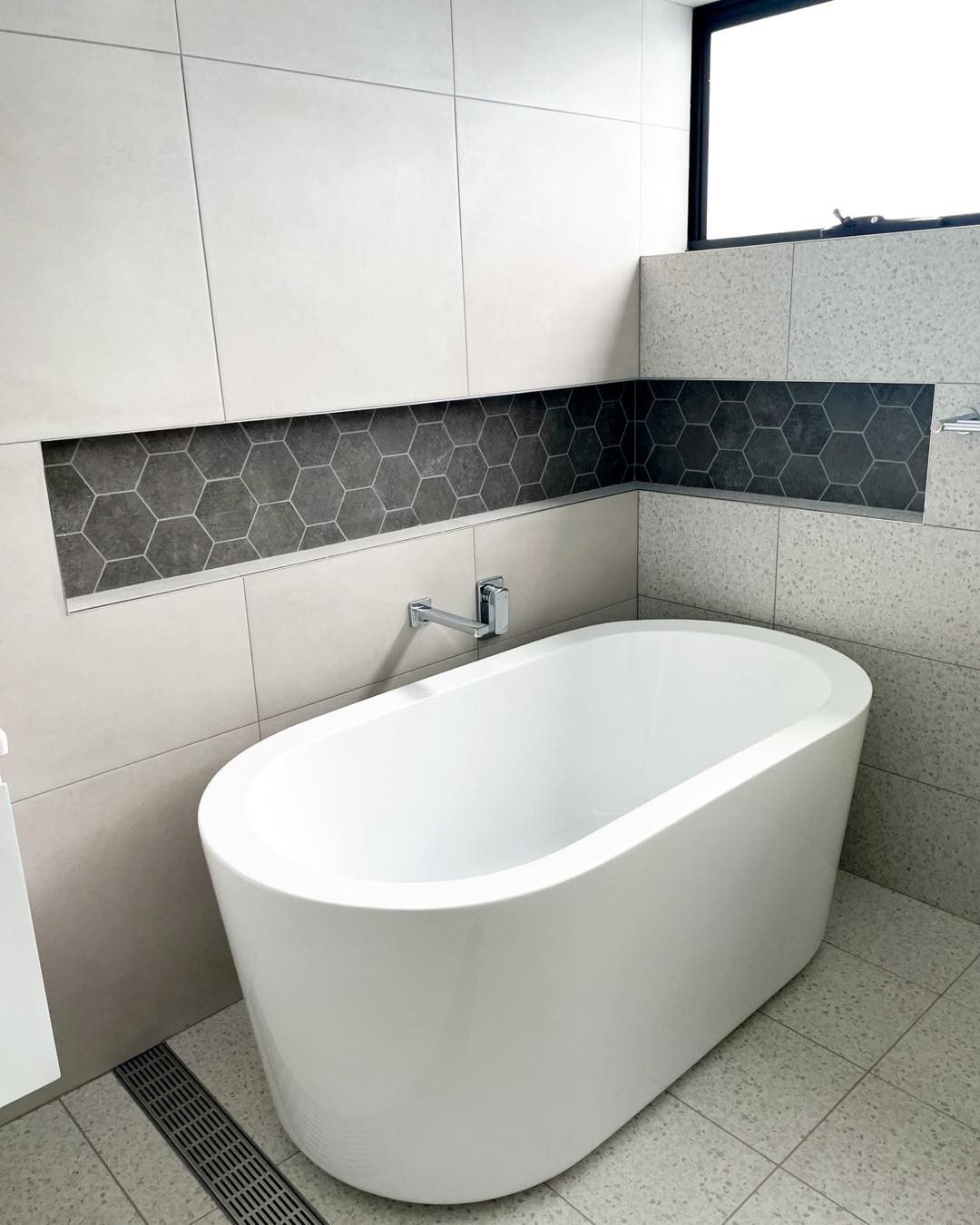 Modern bathroom with a white freestanding tub, gray tiled walls and floor, and a small window.