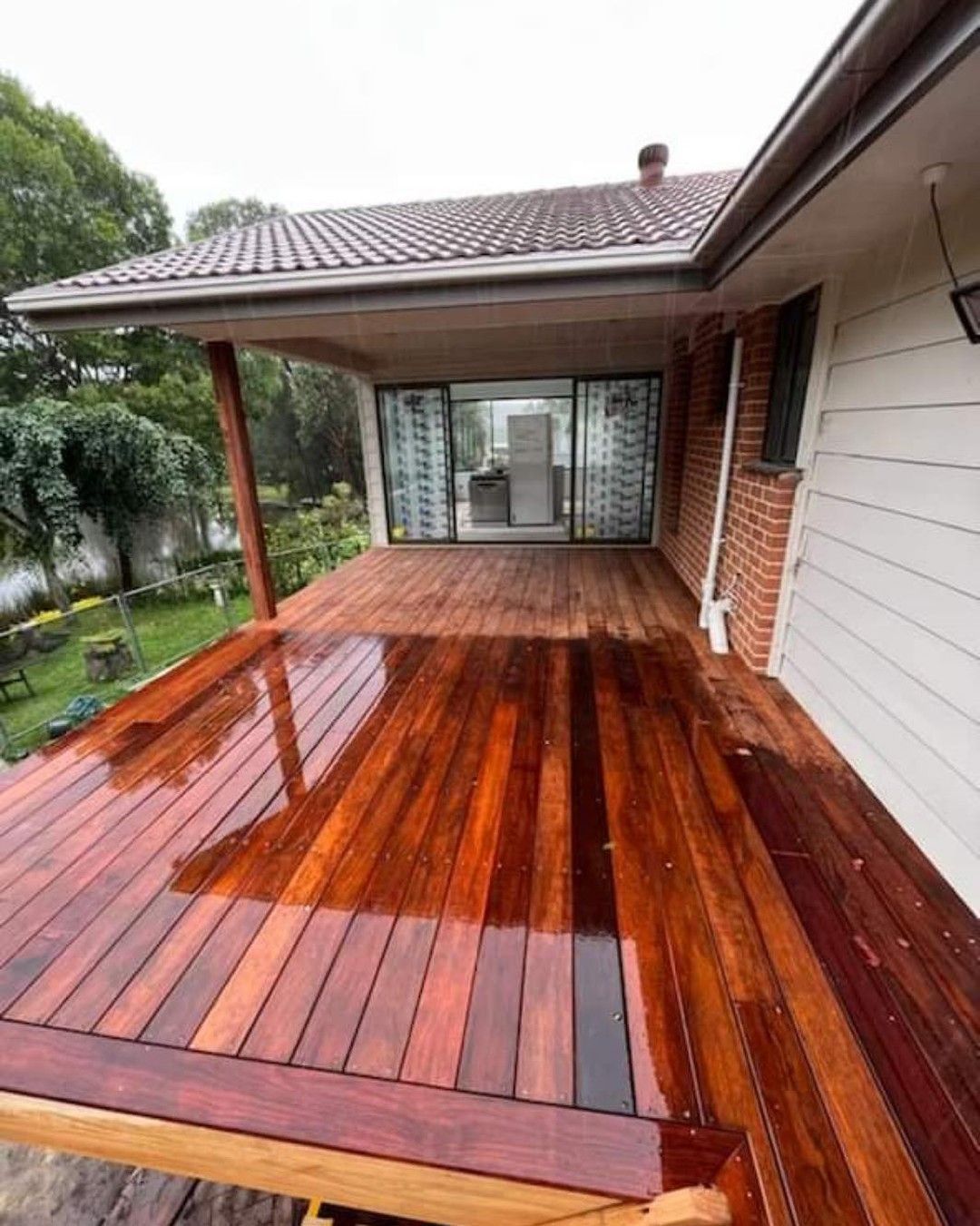 Covered wooden deck with glossy wet boards beside a house and garden view