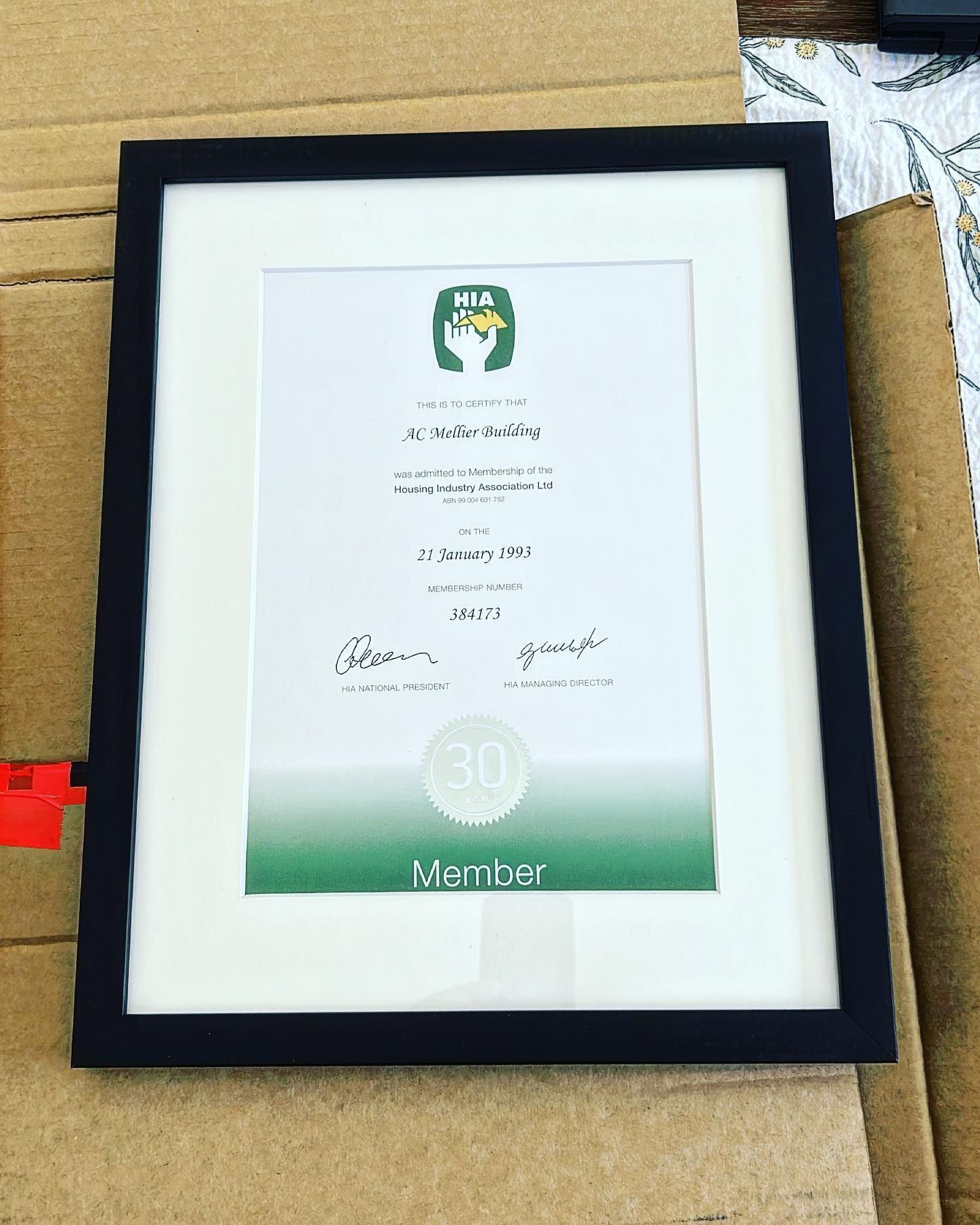 Framed certificate with green gradient accents resting in a cardboard box on a black foam backing.
