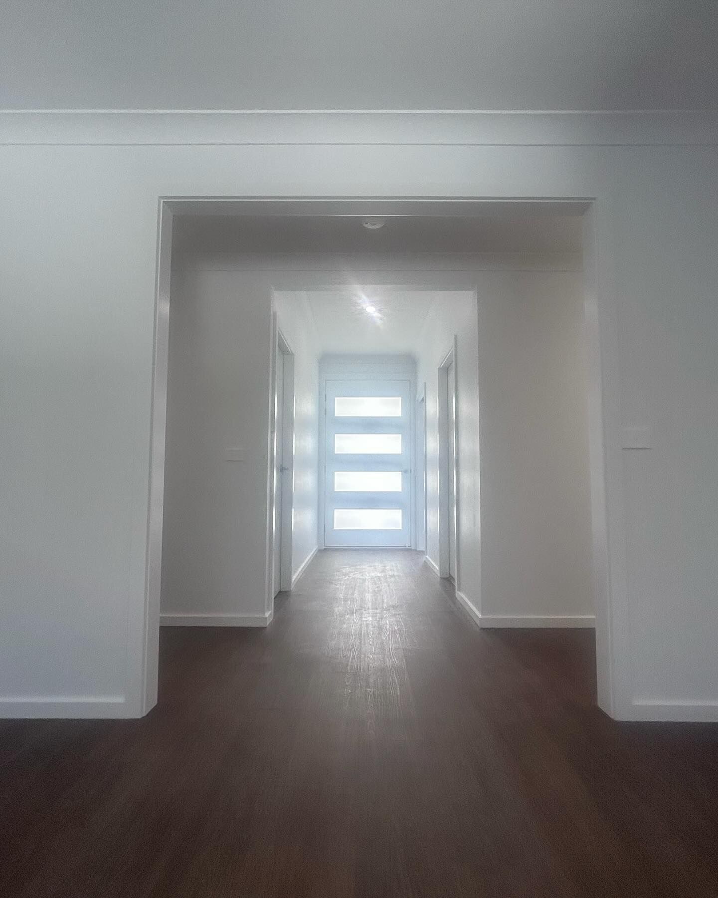 Empty hallway with dark wood floor and white walls leading to a bright glass door.