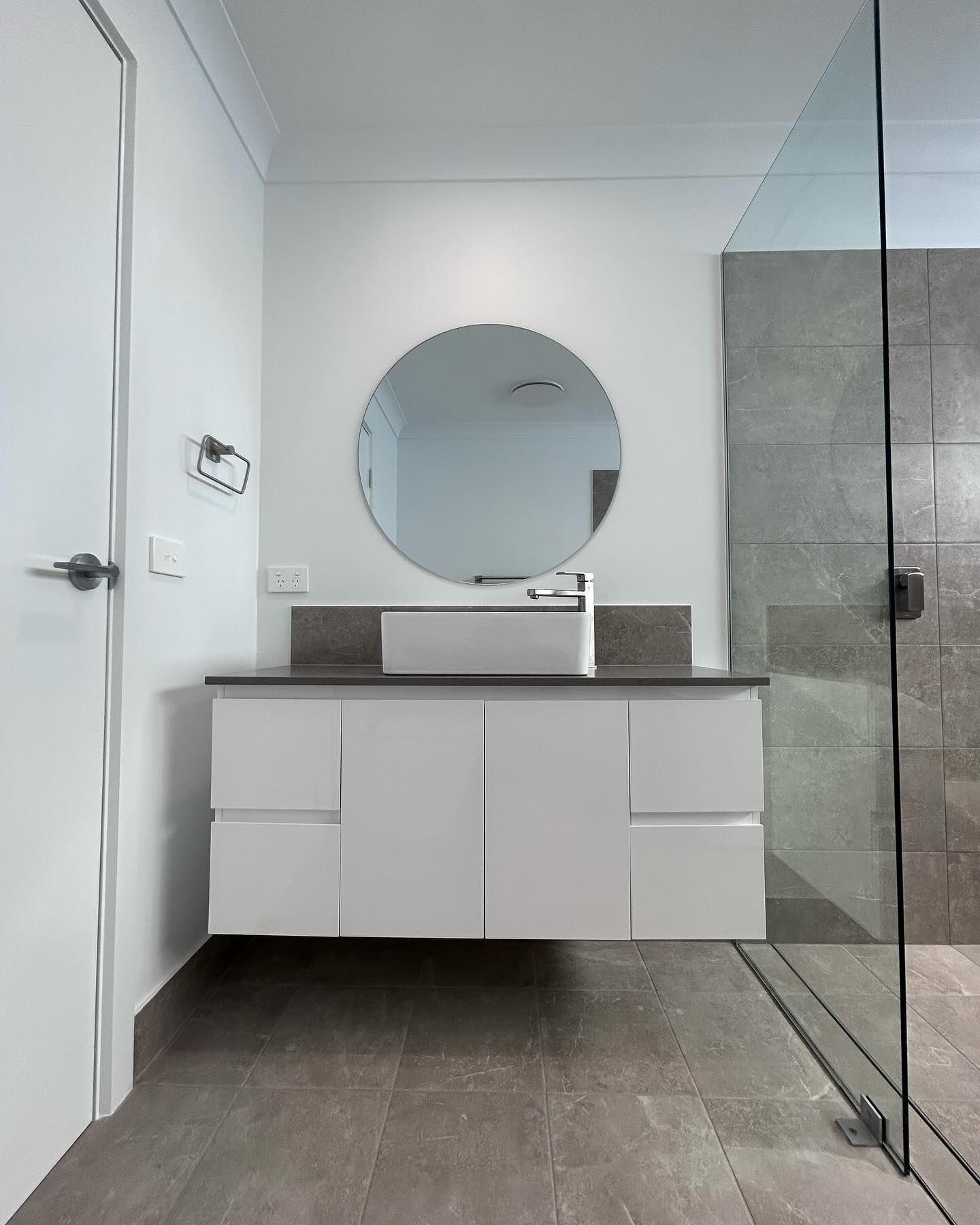 Modern bathroom with white vanity, round mirror, and glass shower enclosure on gray tile floor