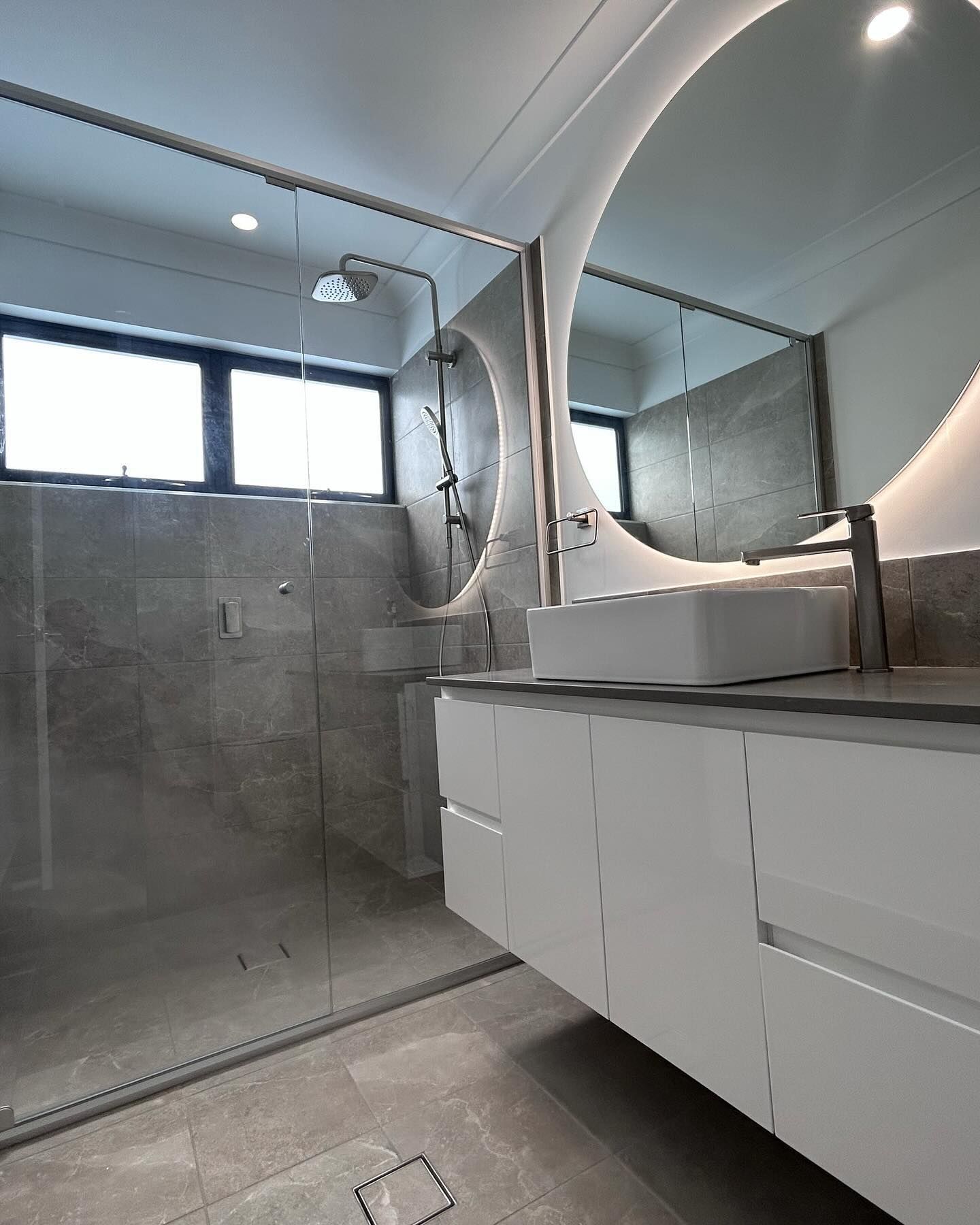 Modern bathroom with glass shower, gray tile, oval mirror, and white vanity sink.