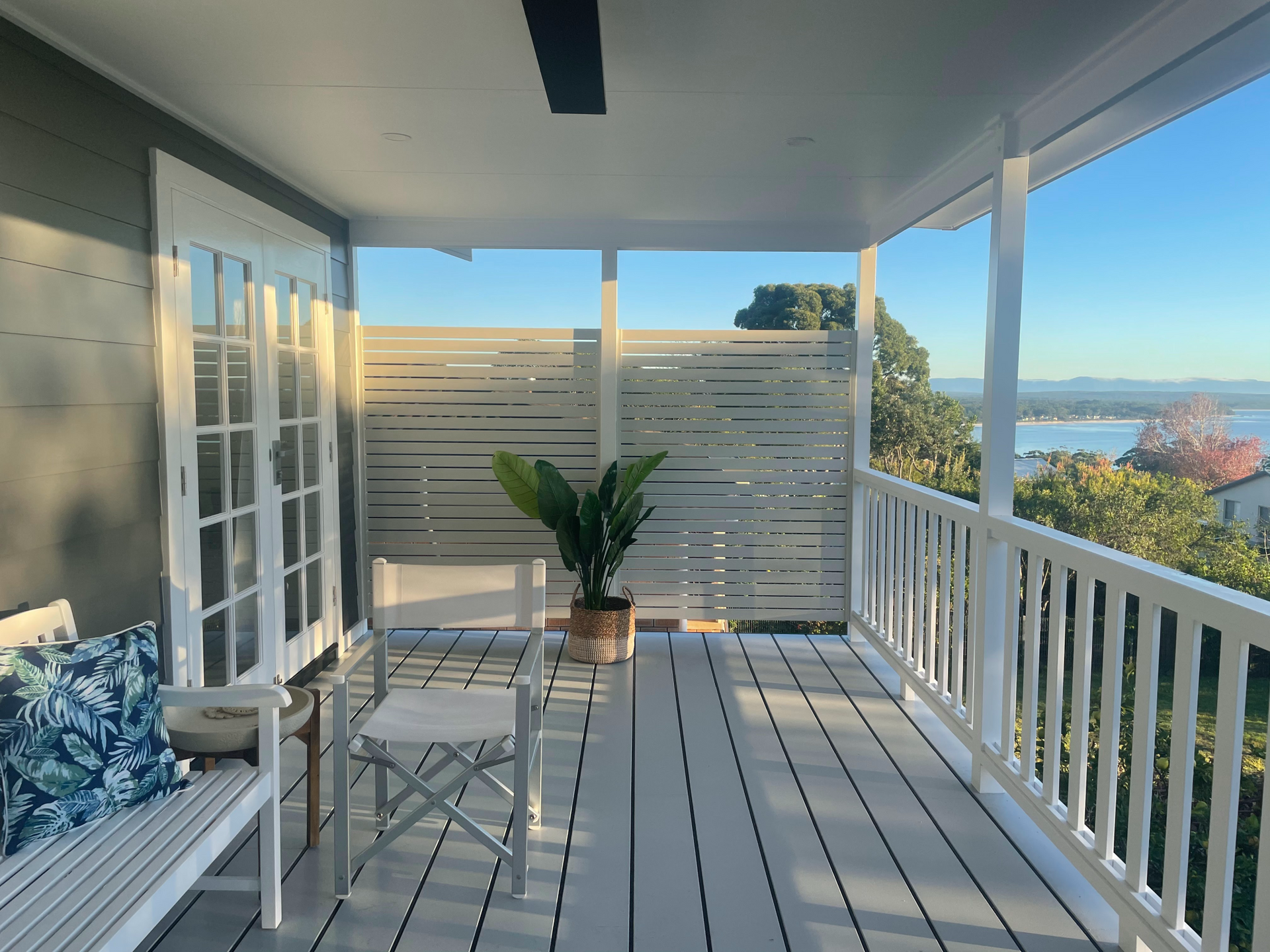 Covered seaside porch with white railing, teal chairs, and potted plant overlooking the water
