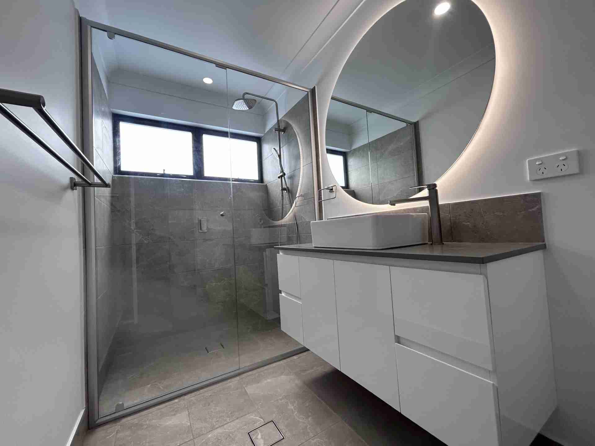 Modern bathroom with glass shower, gray tile, and white vanity with round mirror.