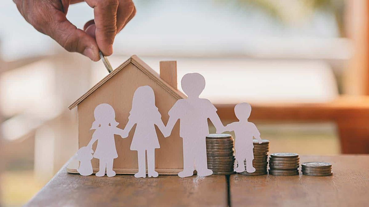 Hands placing a paper family silhouette beside a house model and stacked coins on a table