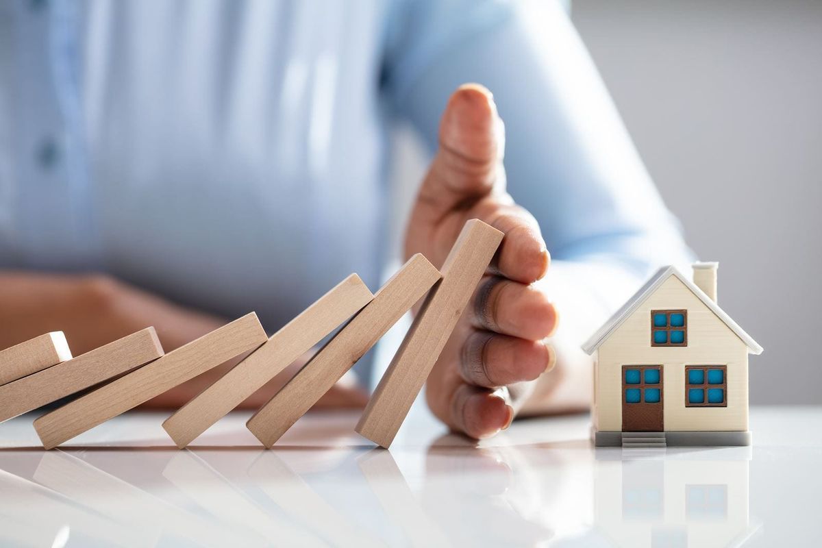 Hand stopping falling dominoes beside a small house model, symbolizing protecting a home from collapse