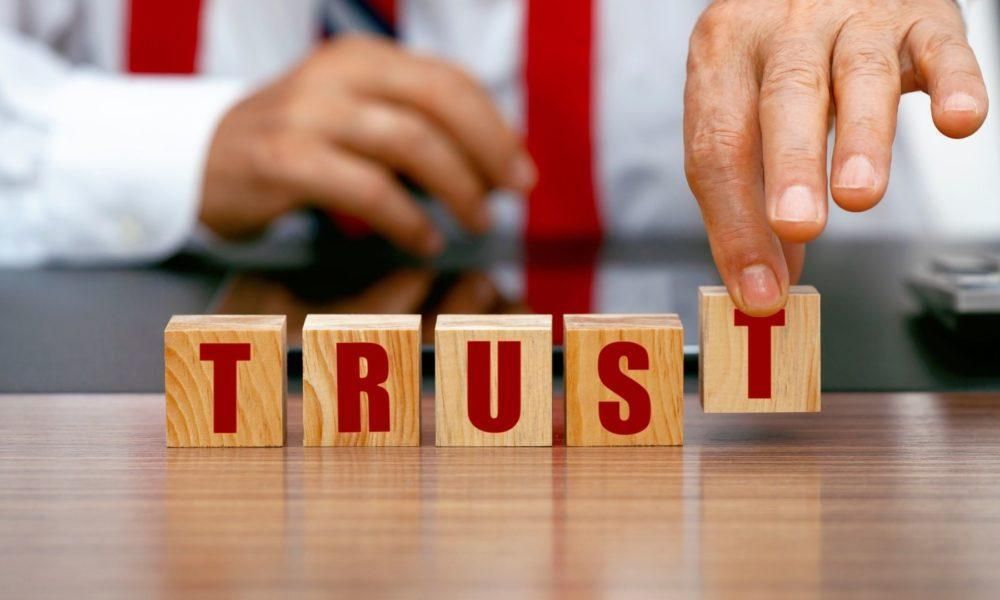 Hands placing a wooden block spelling “TRUST” on a table, with blurred hands in the background.