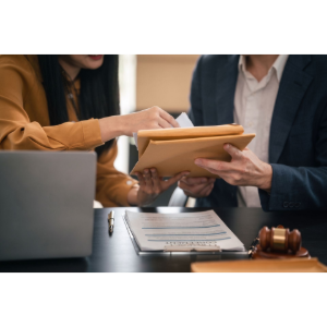 Two people exchanging a folder over a desk with a laptop and gavel in a meeting room