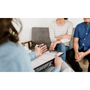 Small group in a counseling session, seated on a couch while a person gestures with a notebook in the foreground