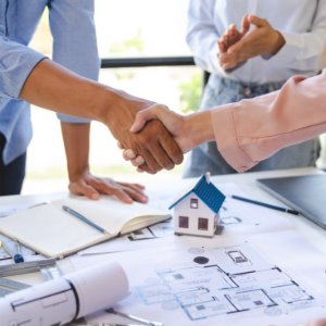 Two people shake hands over house plans and a small model house on a desk