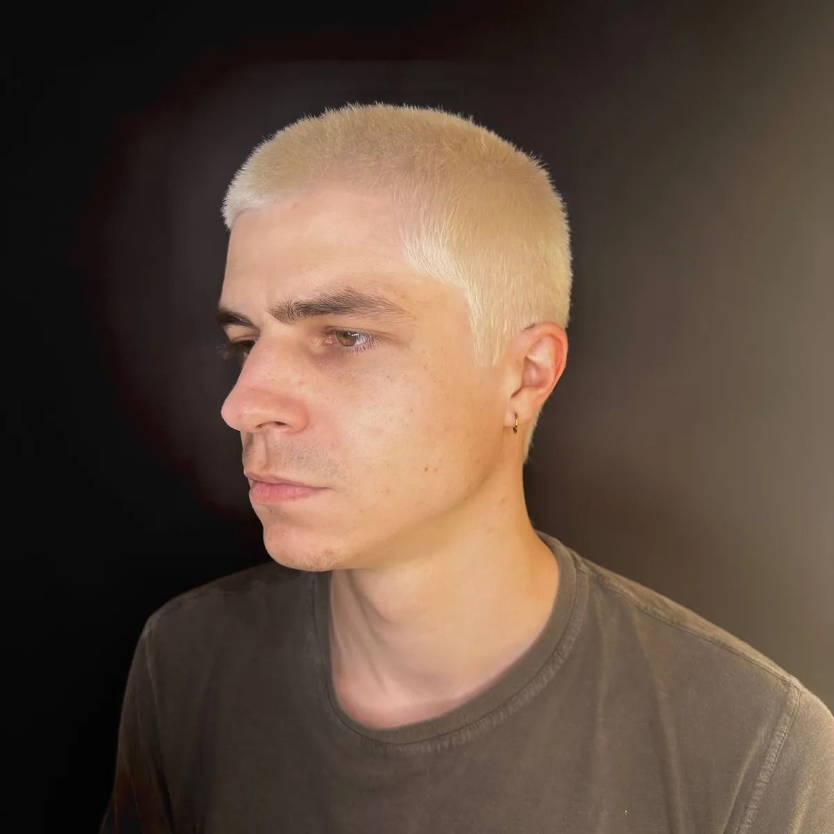 Ritratto di una persona con i capelli corti decolorati, vestita con una maglia scura su sfondo nero