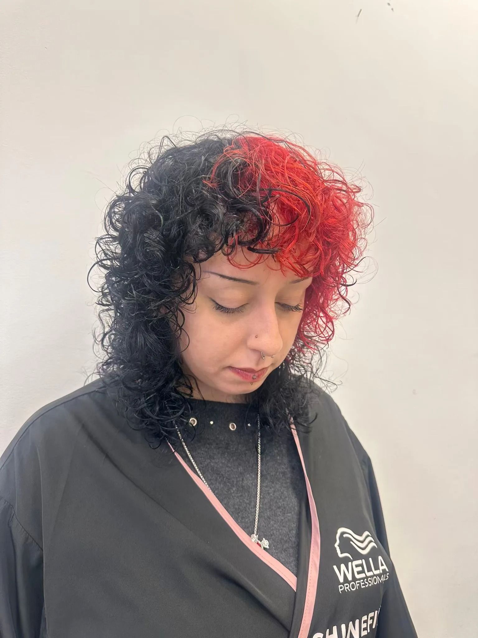 Persona con capelli ricci neri e rossi che guarda verso il basso, con indosso una maglietta nera e una collana, davanti a una parete spoglia.