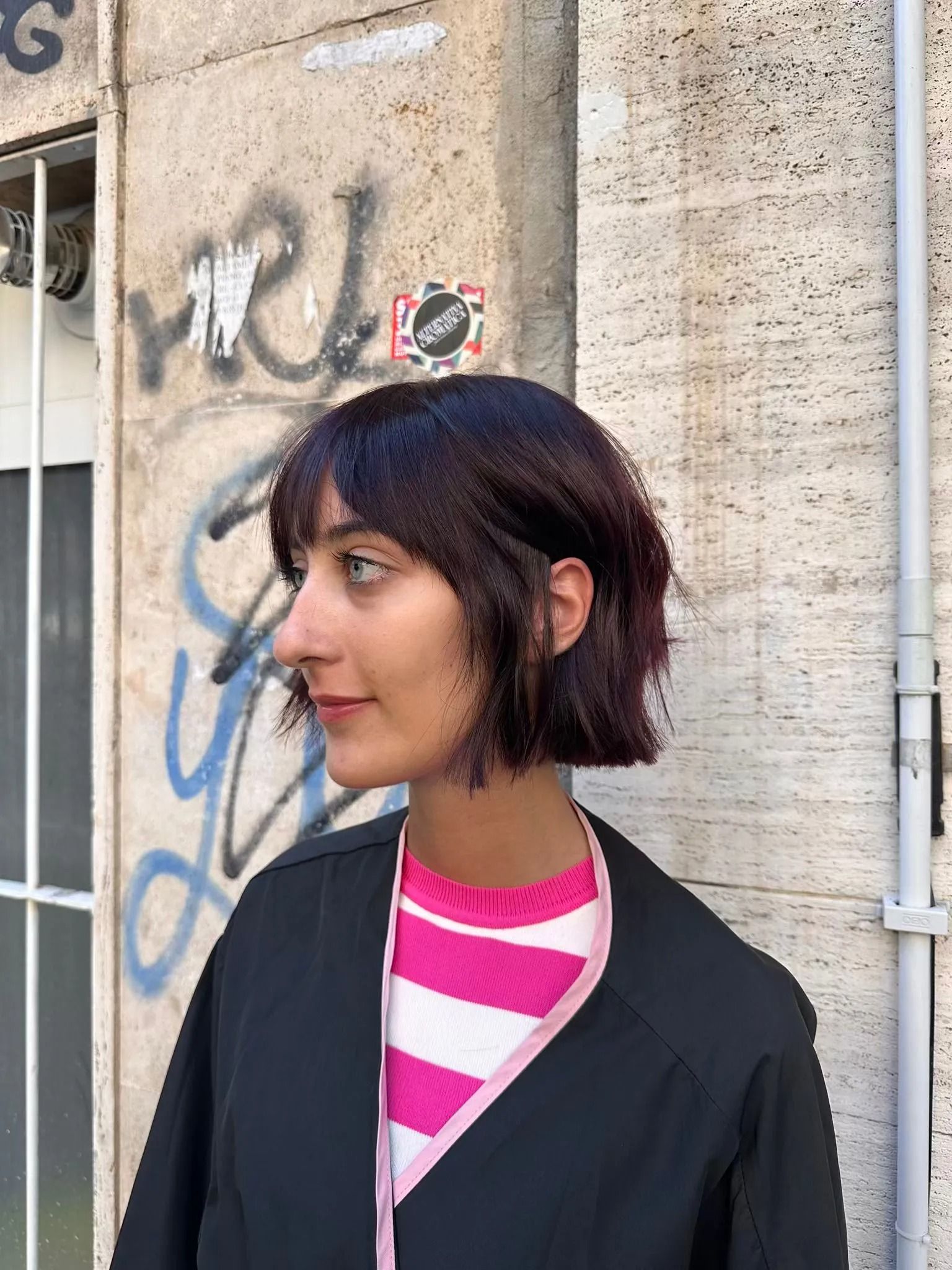 Persona di profilo con una giacca nera e una camicia a righe rosa, davanti a un muro coperto di graffiti.