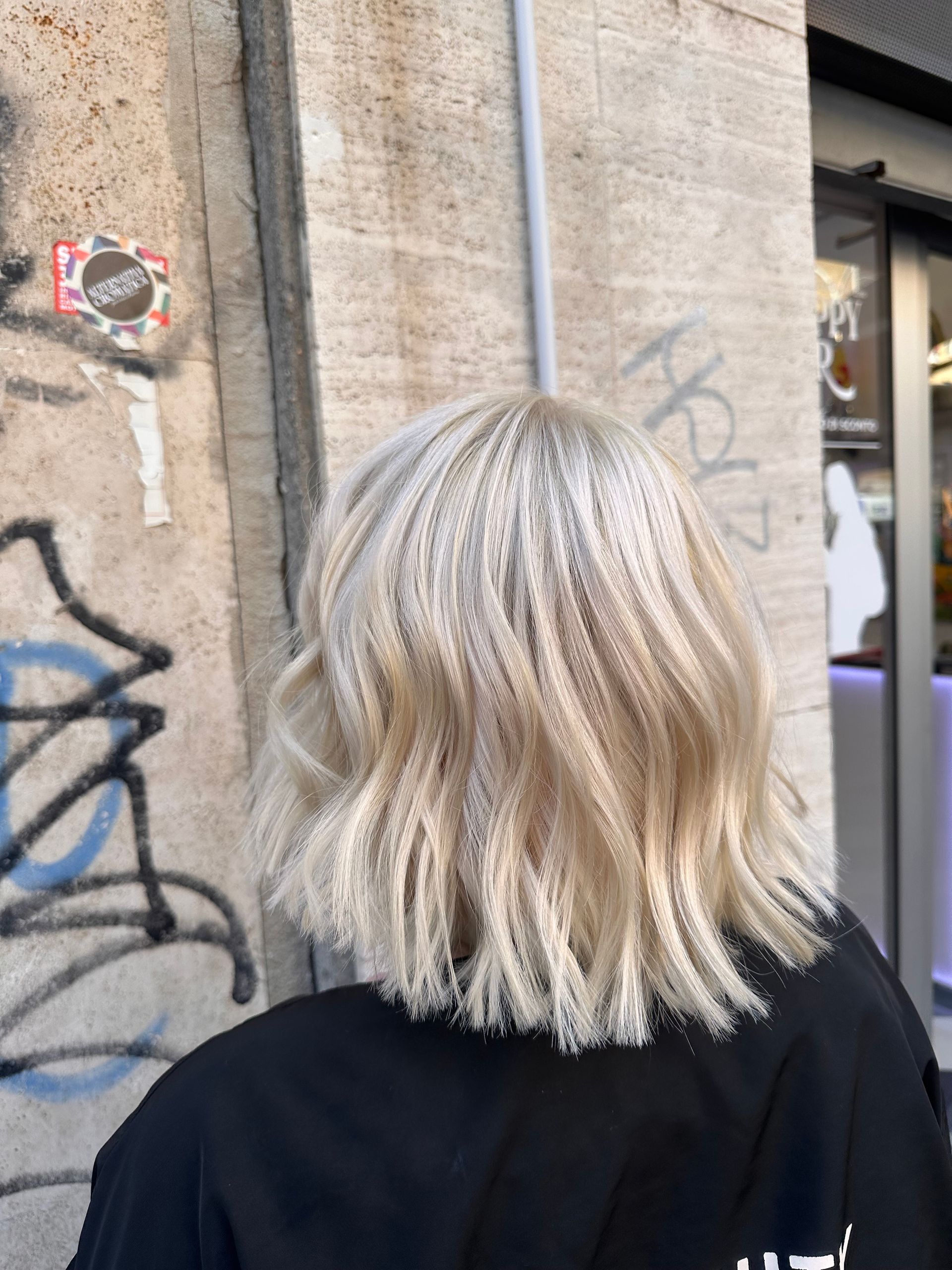 Vista da dietro di una persona con capelli biondo platino mossi, che indossa una giacca nera e sta accanto a un muro ricoperto di graffiti