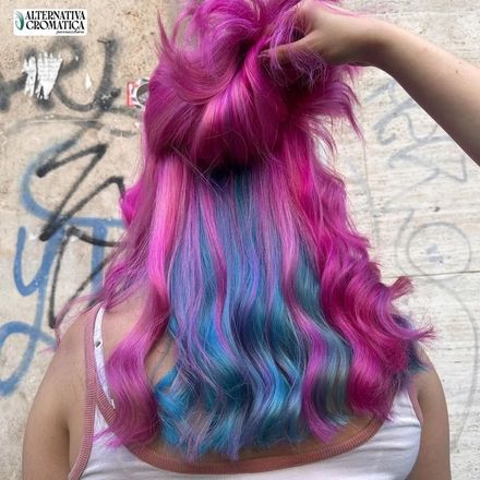 Persona con lunghi capelli magenta, blu e viola, vista di spalle davanti a un muro ricoperto di graffiti.