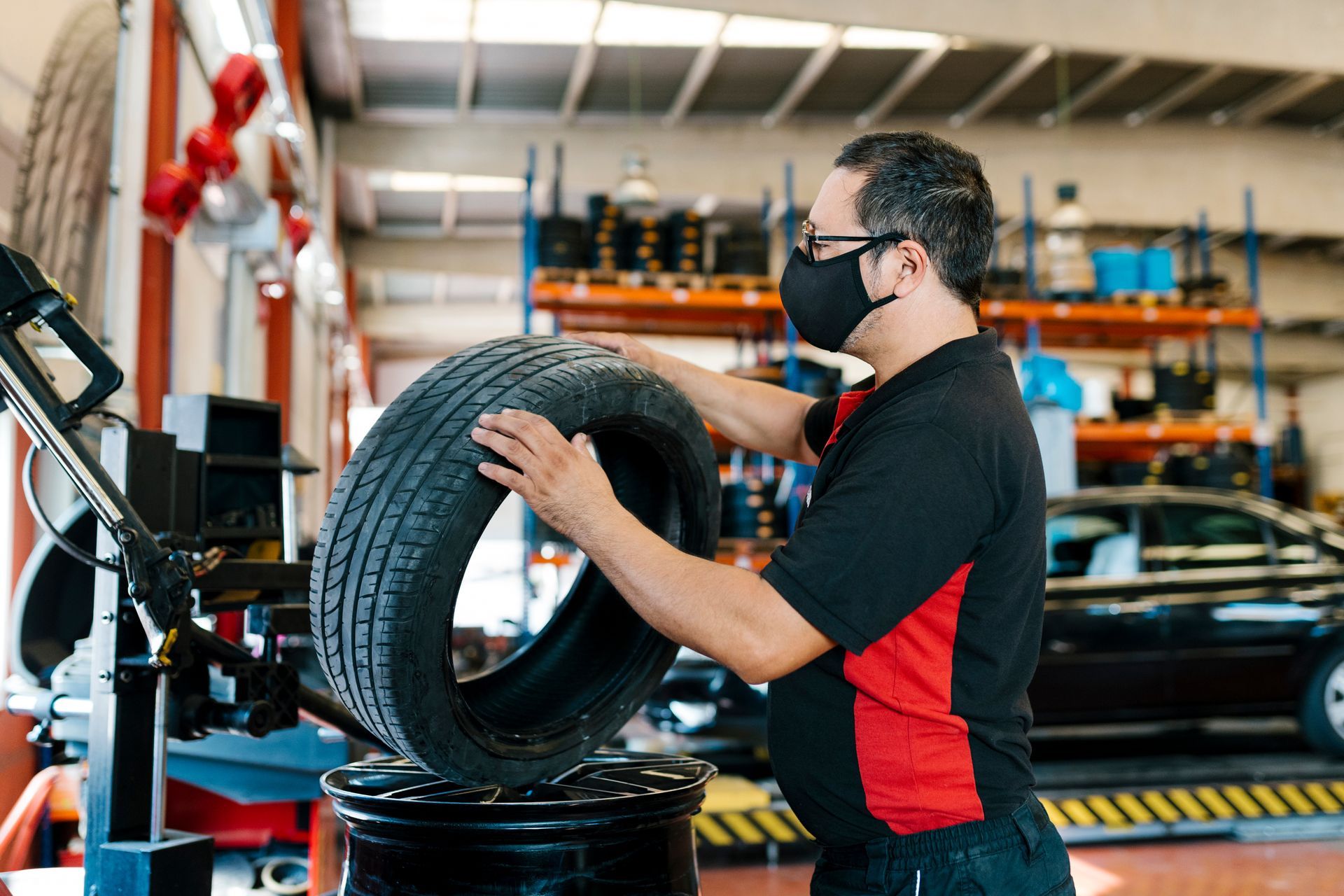 A mechanic wearing a mask is examining a tire in an auto repair shop. A mechanic wearing a mask is examining a tire in an auto repair shop.