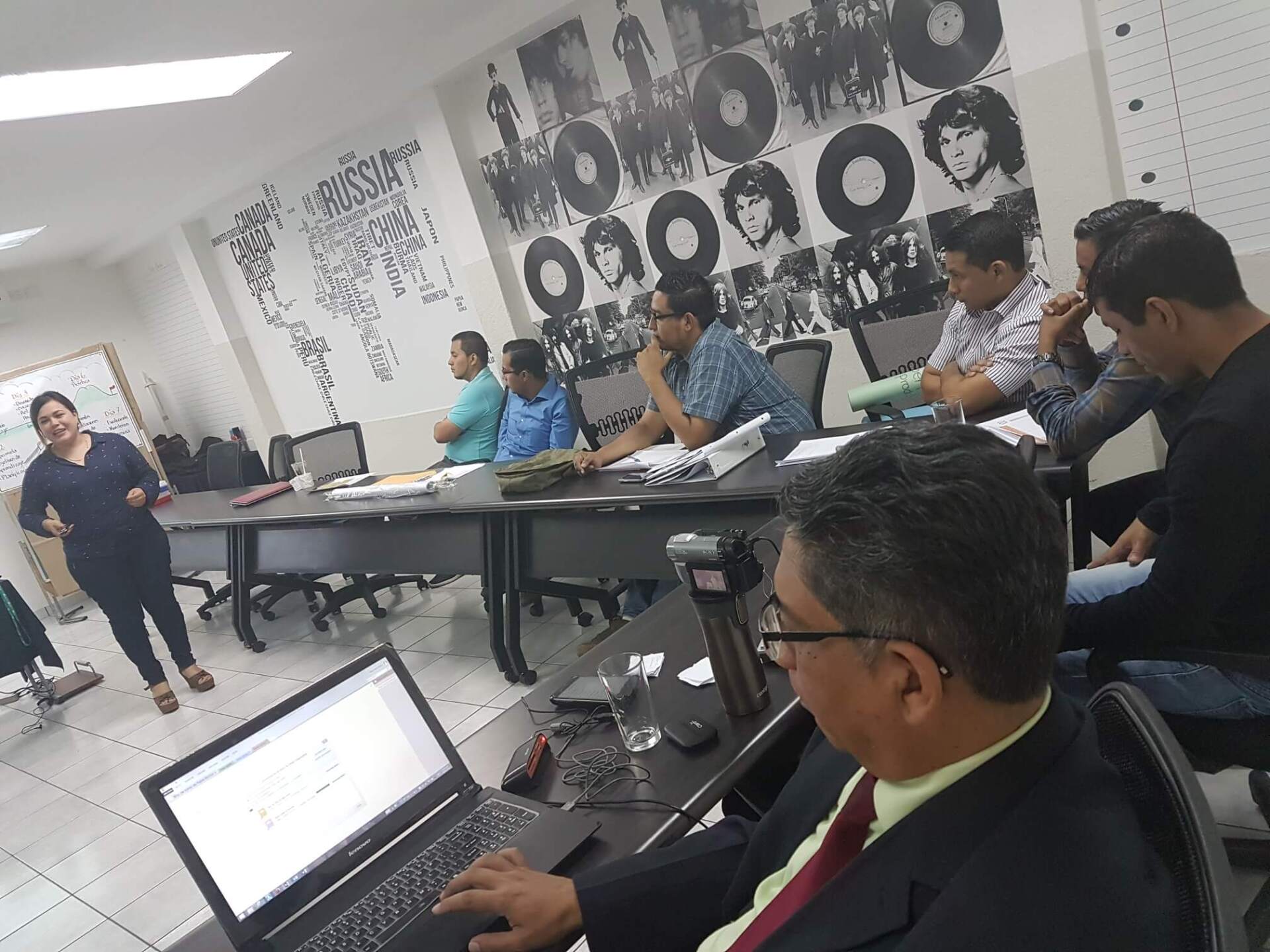 Reunión en una sala de conferencias con personas sentadas alrededor de una mesa, una computadora portátil abierta en primer plano y una obra de arte en la pared detrás de ellas.