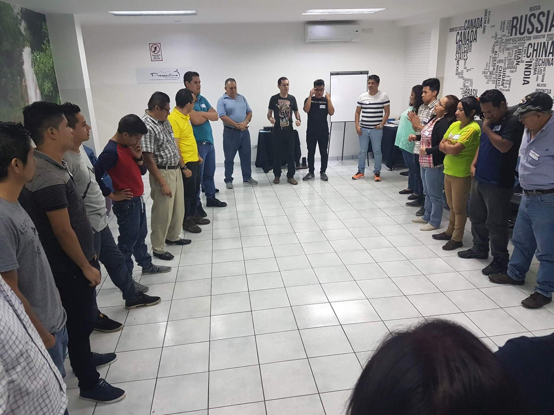 Varias personas de pie formando un semicírculo en una habitación alicatada, frente a un orador situado en el centro.