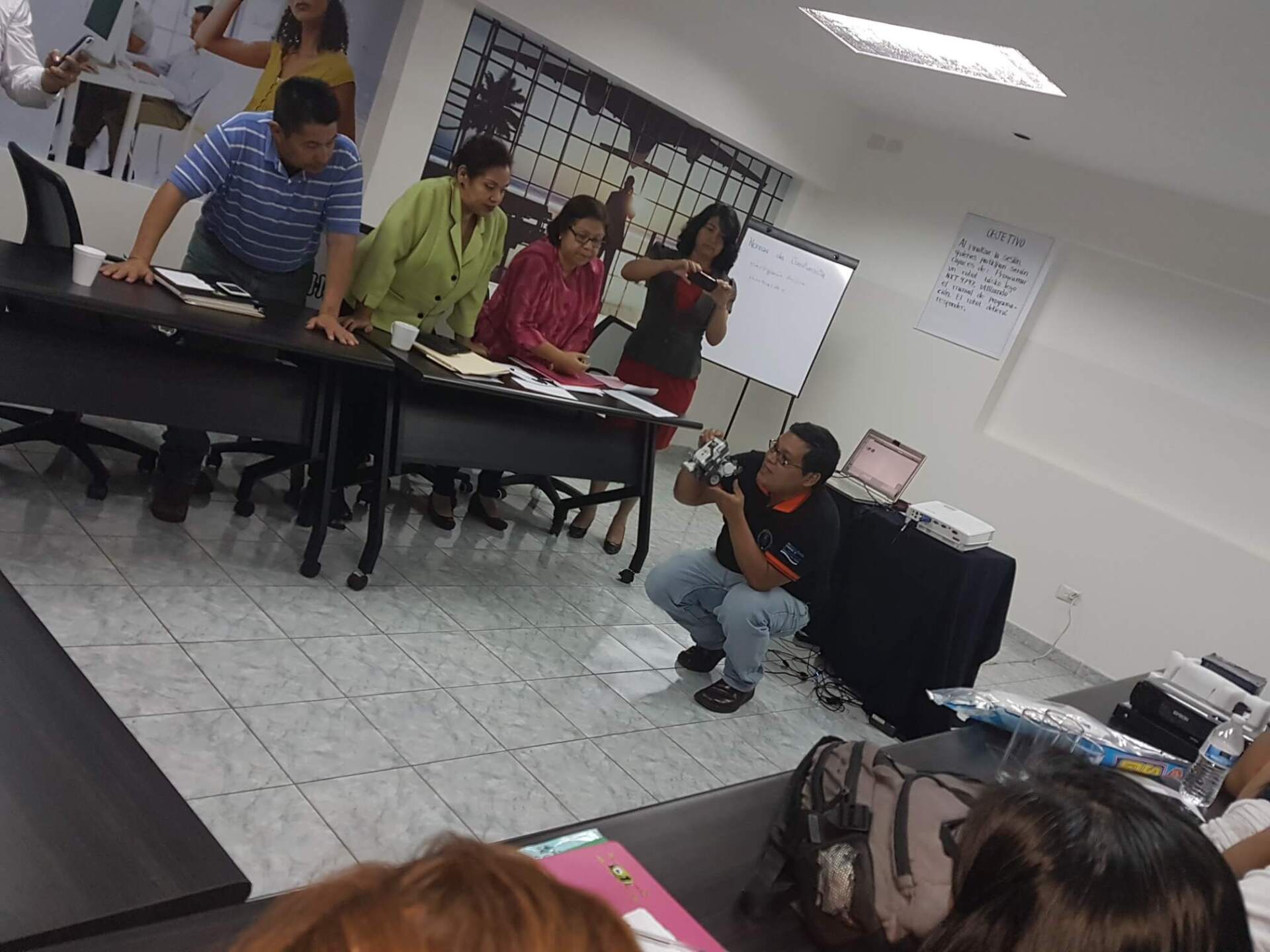Varias personas se reunieron en un aula, sentadas y de pie alrededor de las mesas, y una de ellas se agachó para tomar una foto.