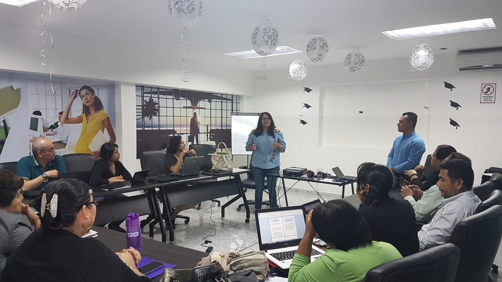 Reunión en una oficina luminosa con gente sentada alrededor de mesas mientras una persona realiza la presentación al frente.